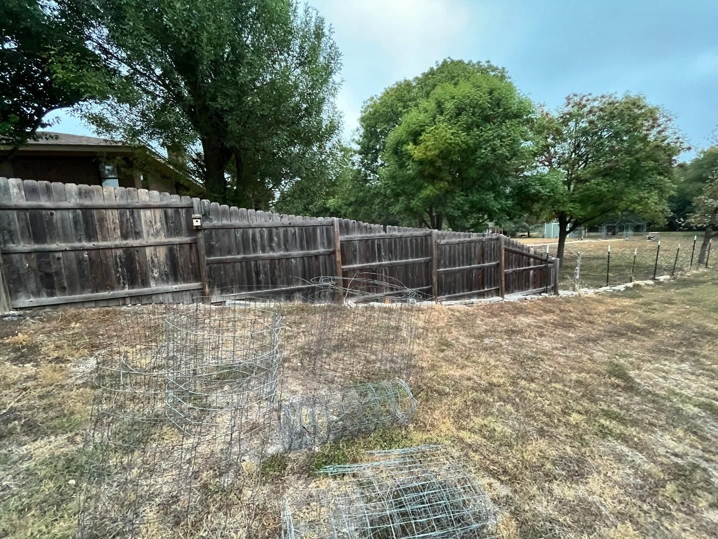 A weathered wooden fence borders a yard with dry grass, trees, and cloudy sky.