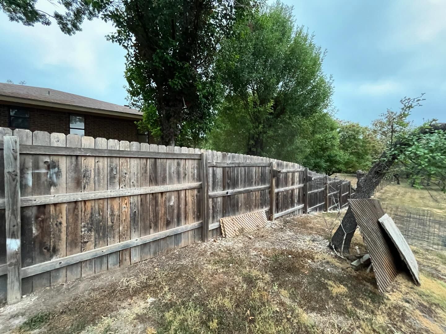 Wooden fence in a yard with dry grass, weathered wood, and green trees against a cloudy sky.