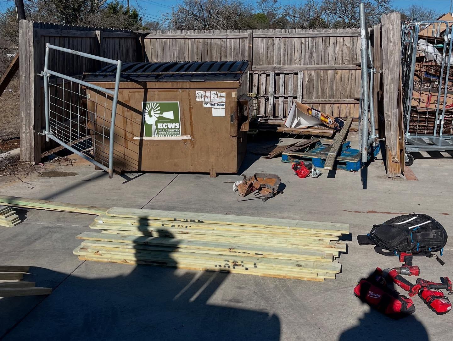 Lumber and tools beside a dumpster and fence outdoors.