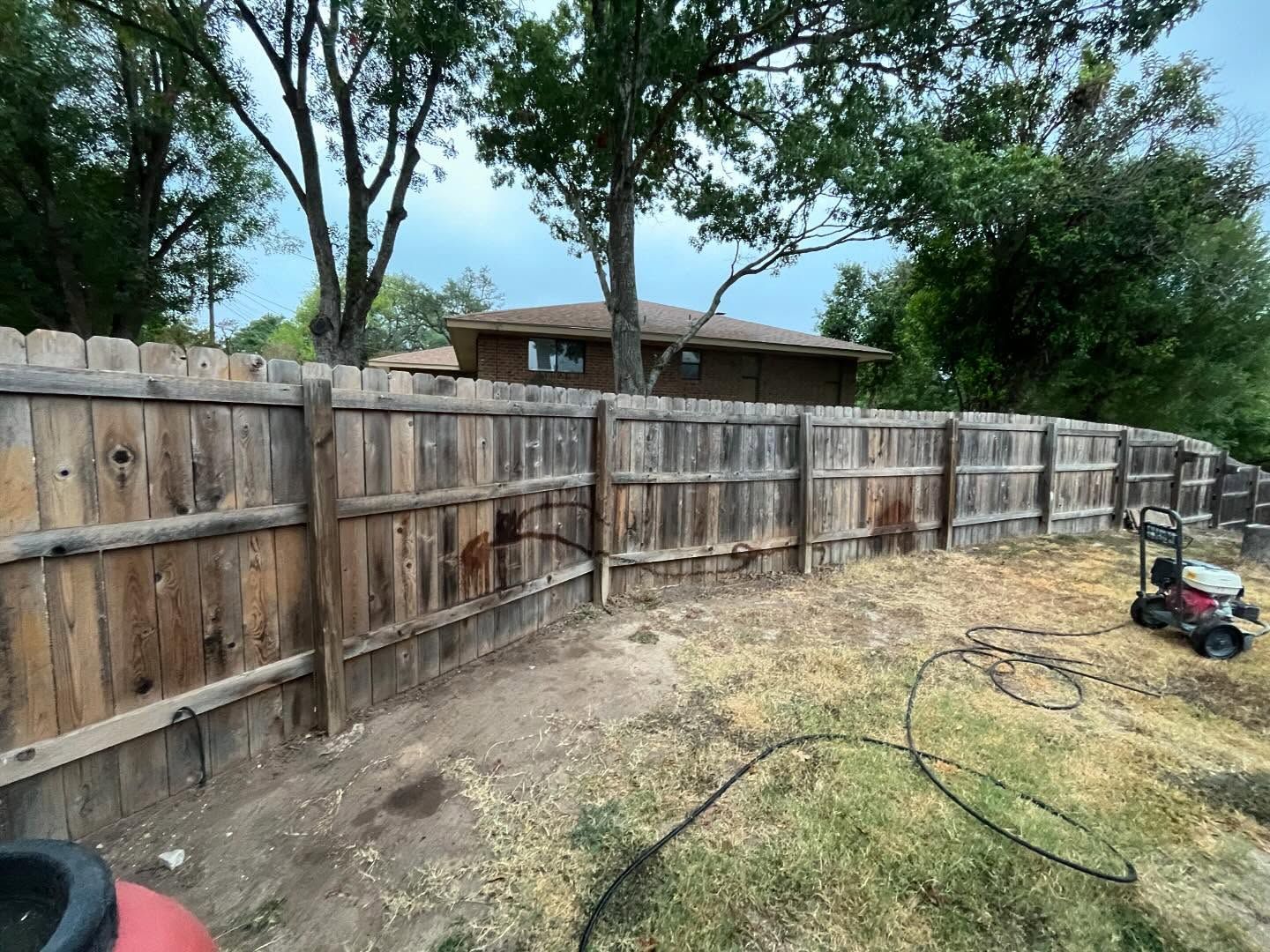 Wooden fence in a backyard with a lawnmower, hose, and a house in the background.