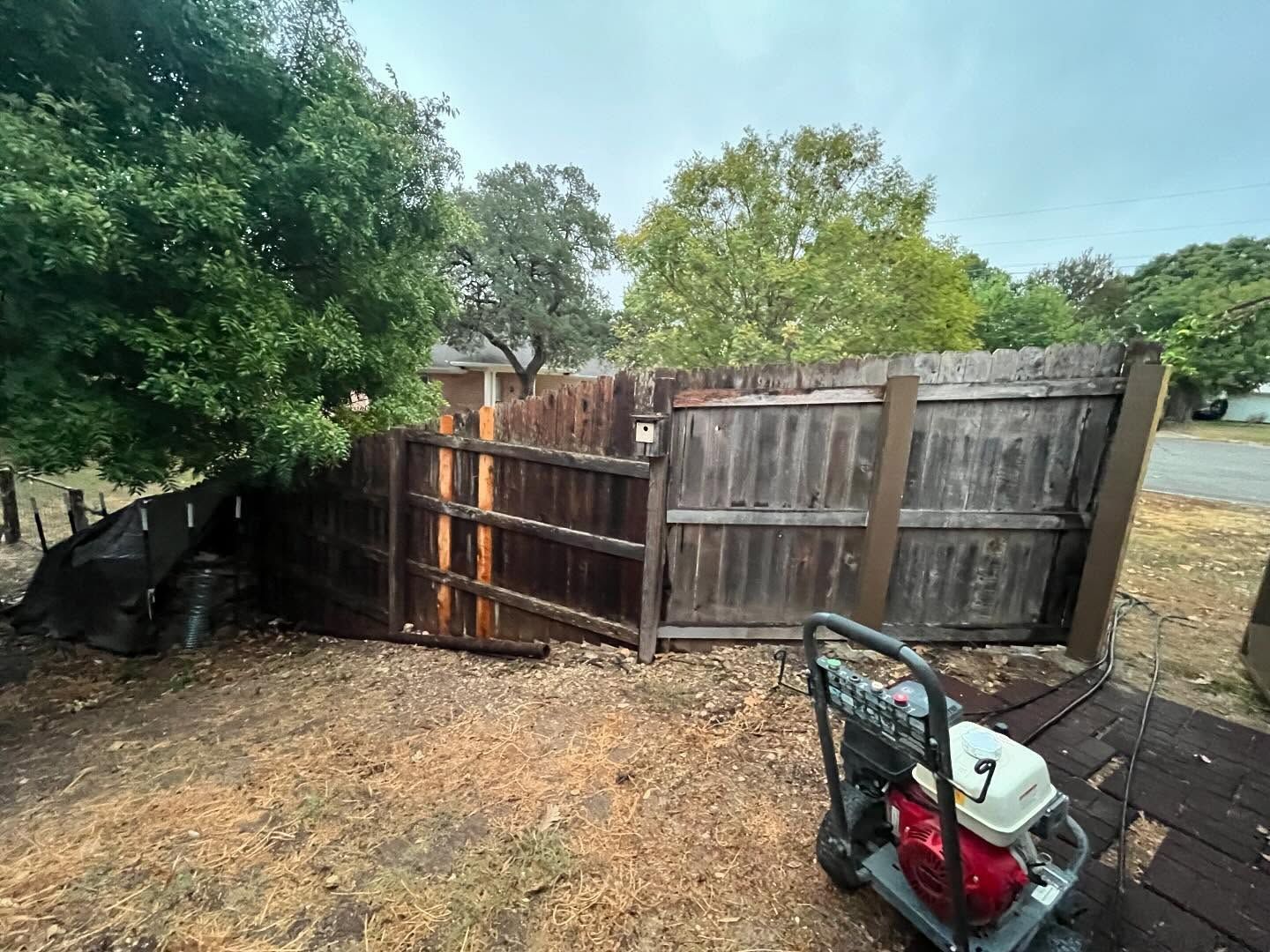 A pressure washer next to a wooden fence. Some fence panels are darker, presumably recently cleaned. Cloudy sky overhead.