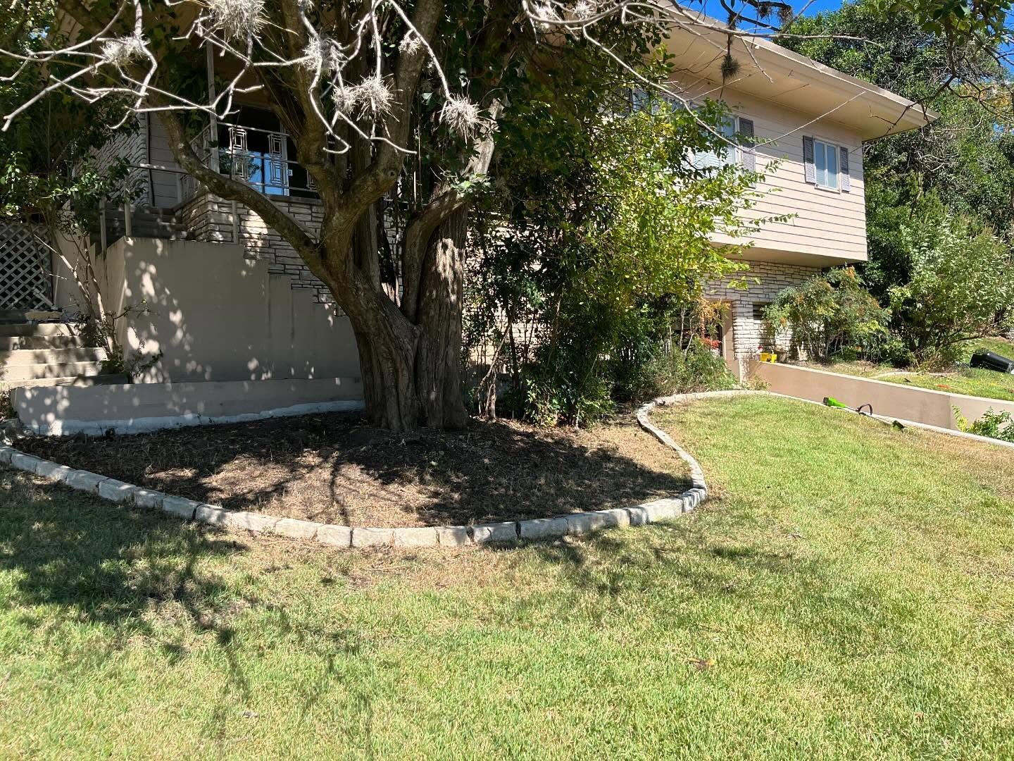Two-story beige house with a large tree in front. Green grass and a paved border around the tree.