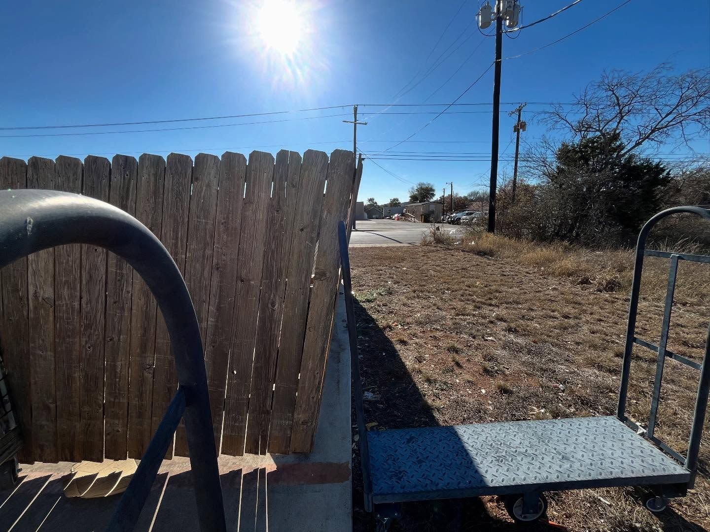 Sun shining over a wooden fence and a hand truck on a sidewalk next to dry grass.