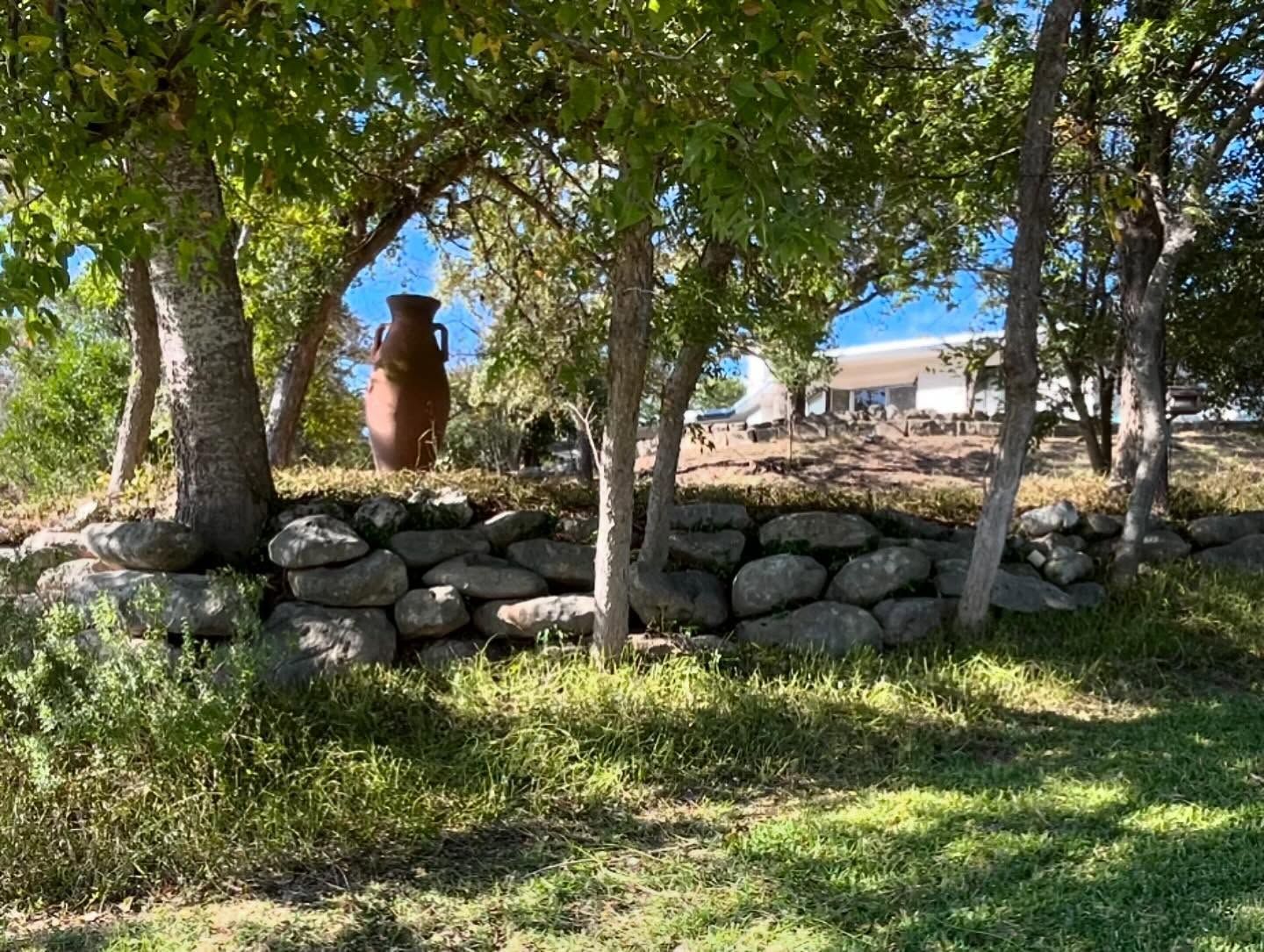 Stone wall supports a garden with trees, a large brown pot, and a distant white building.