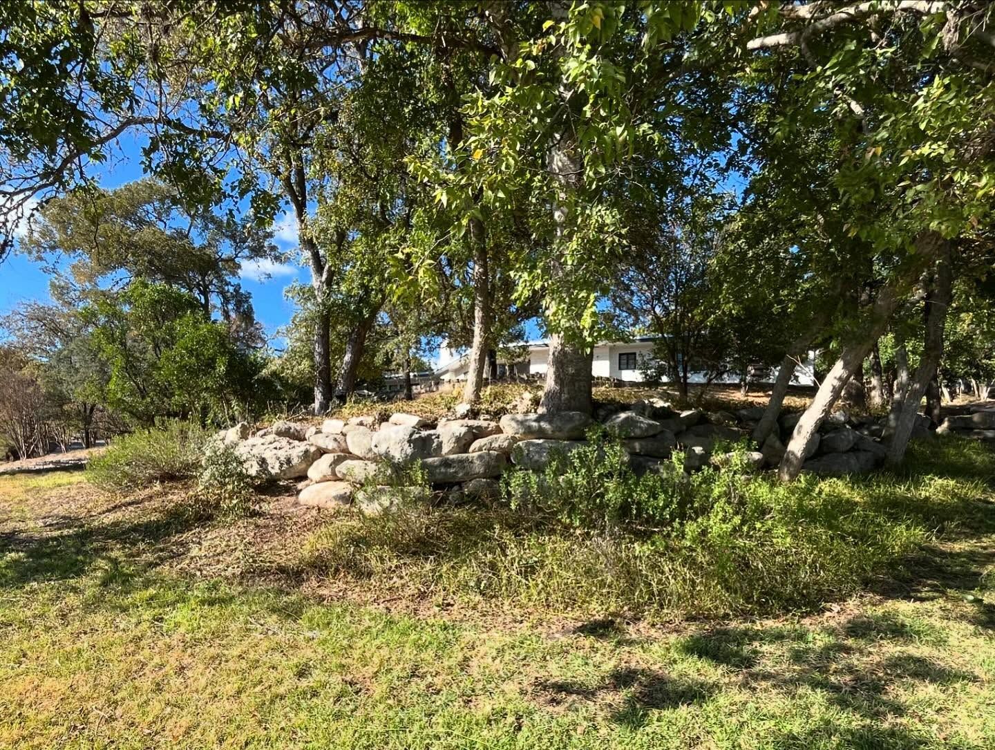 Grassy landscape with large rocks, trees, and glimpses of a white building under a blue sky.