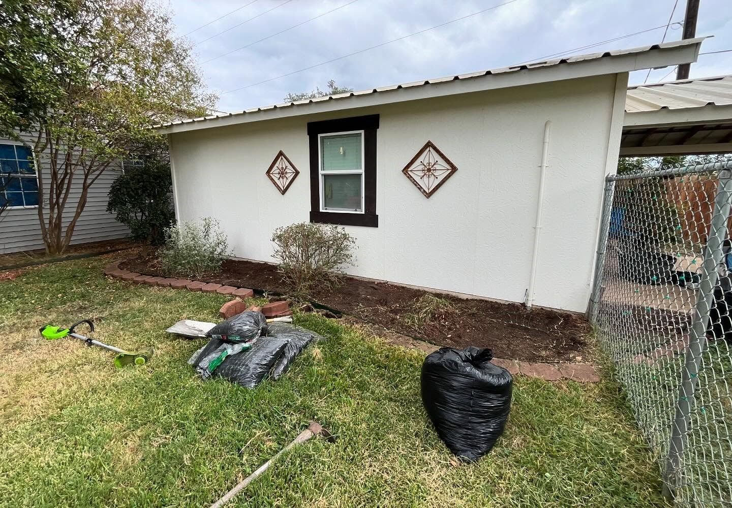 White building with a window and decorative tiles. Yard with grass, mulch, and yard waste bags.