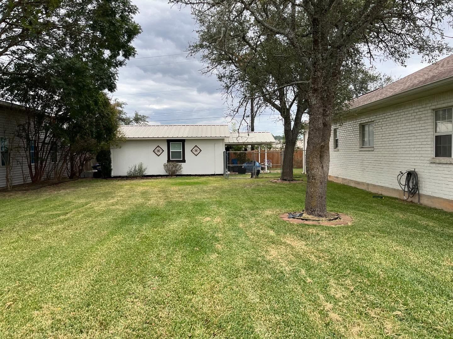 A backyard with green grass, a white building, and trees under a cloudy sky.
