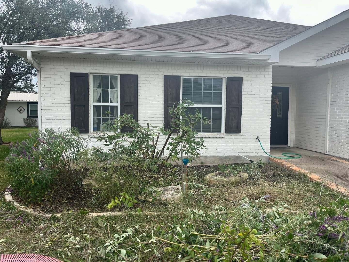 White brick house with brown shutters and a brown roof, overgrown landscaping in front.