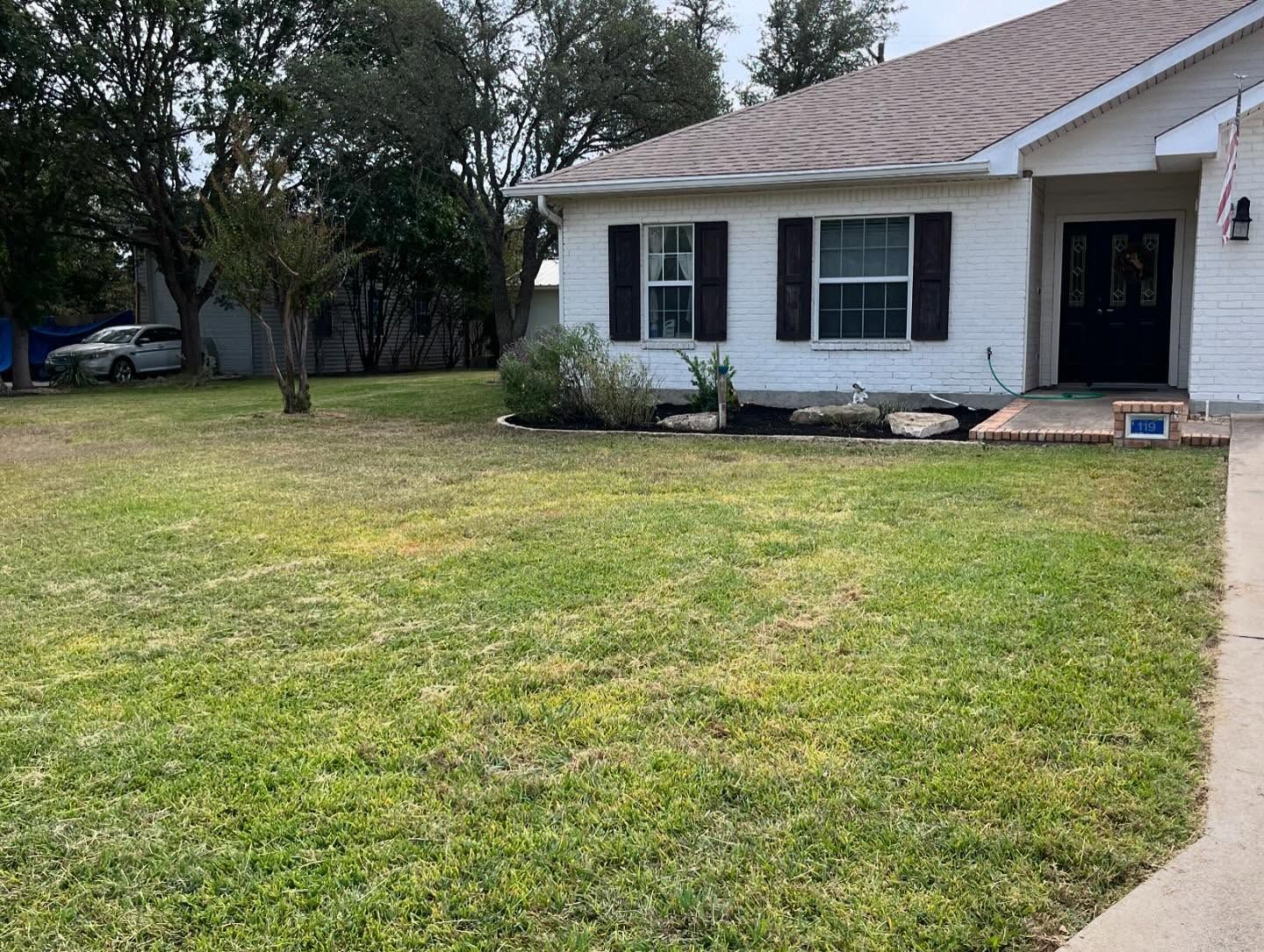 White house with black shutters and door, small front lawn with a car parked to the side, cloudy sky.