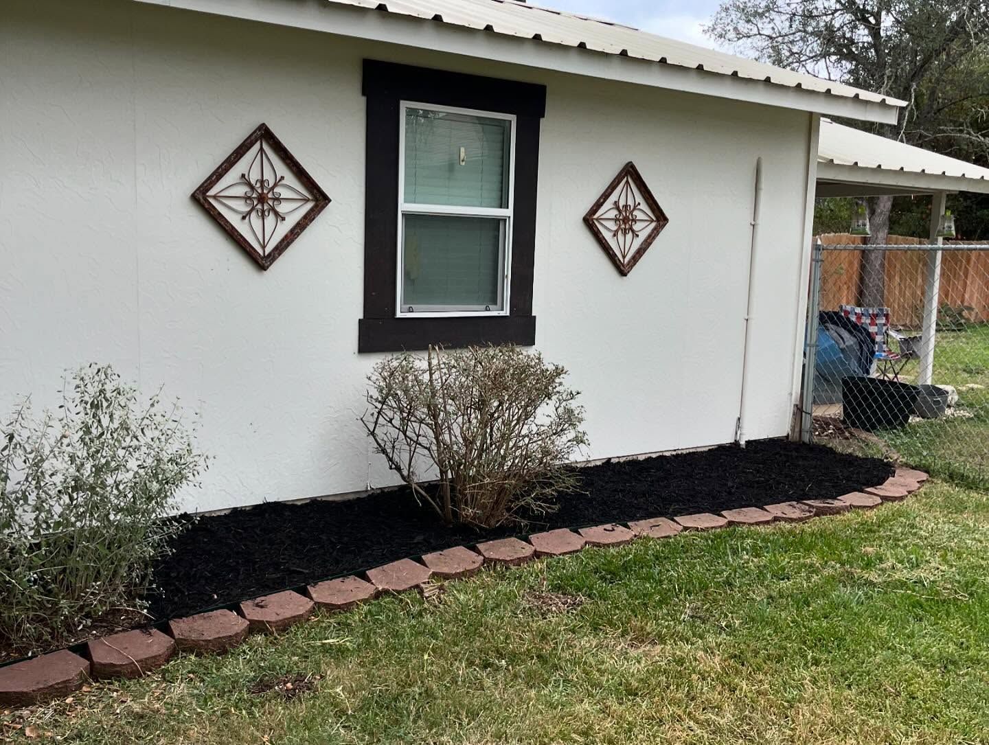 White house exterior with dark brown window trim, two diamond-shaped wall decorations, and a black mulch flower bed with brick edging.