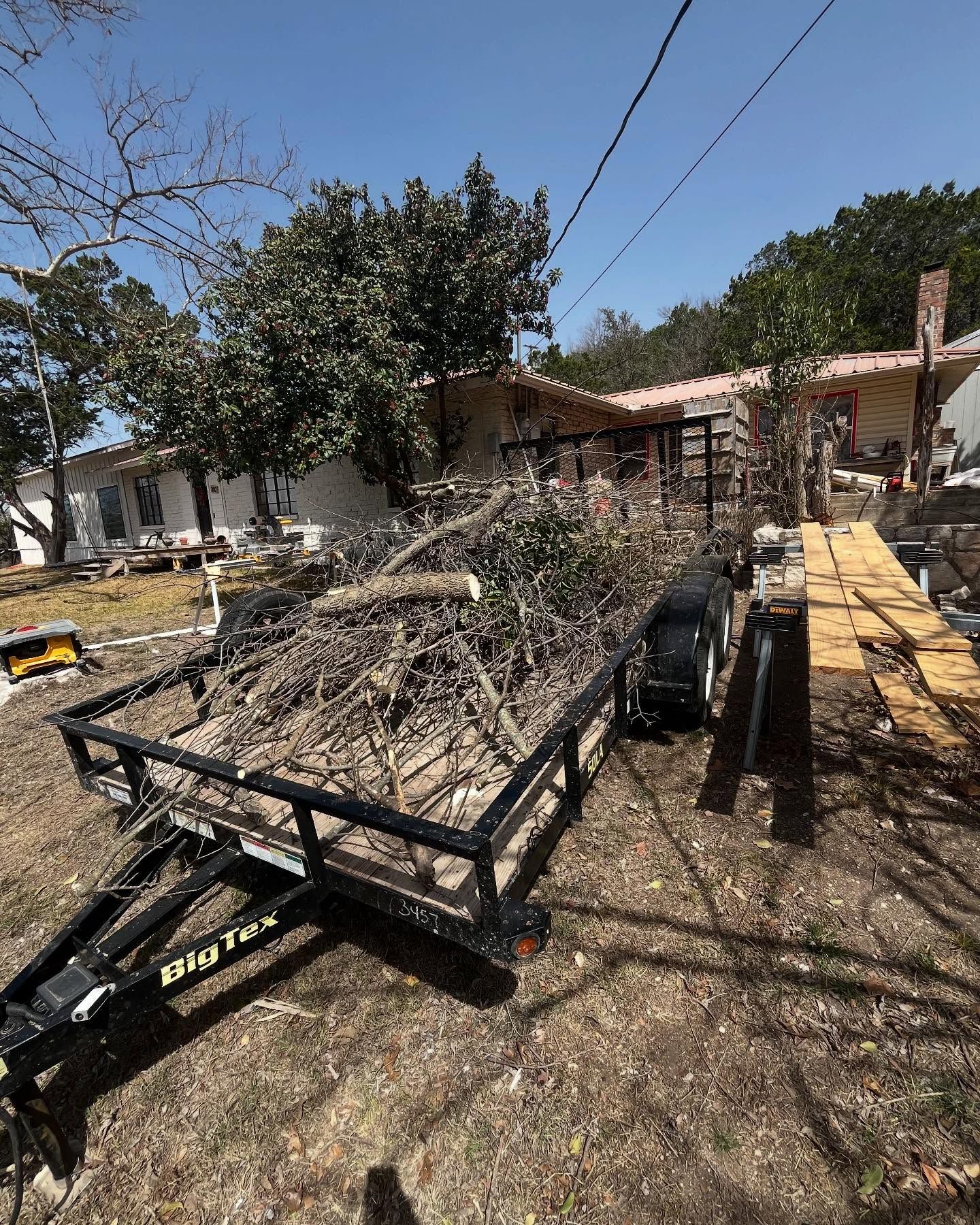 Trailer loaded with branches, parked on dry grass near a house and wooden walkway under a blue sky.