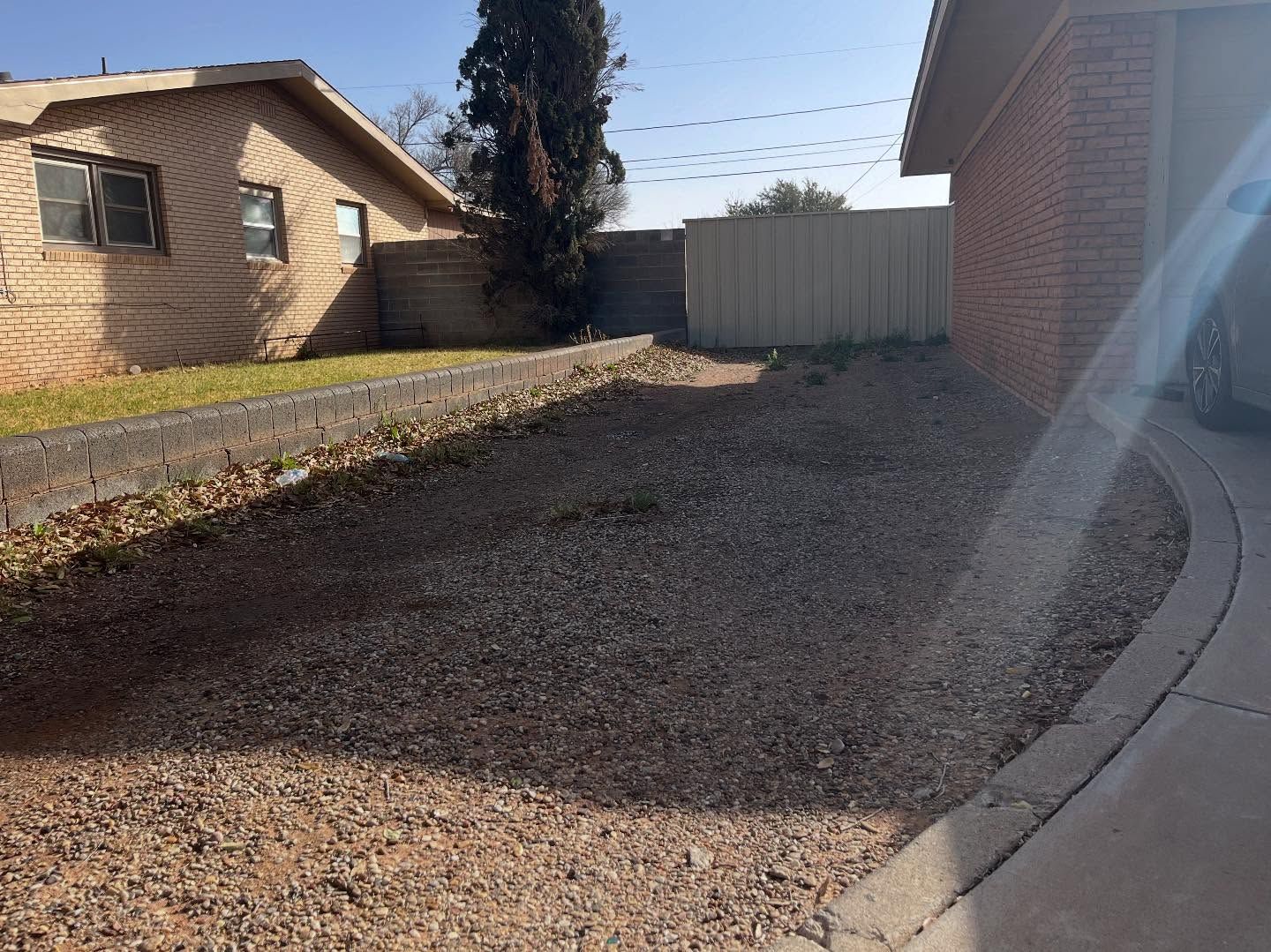 A gravel yard between two brick houses, a curb and a fence, under a blue sky.