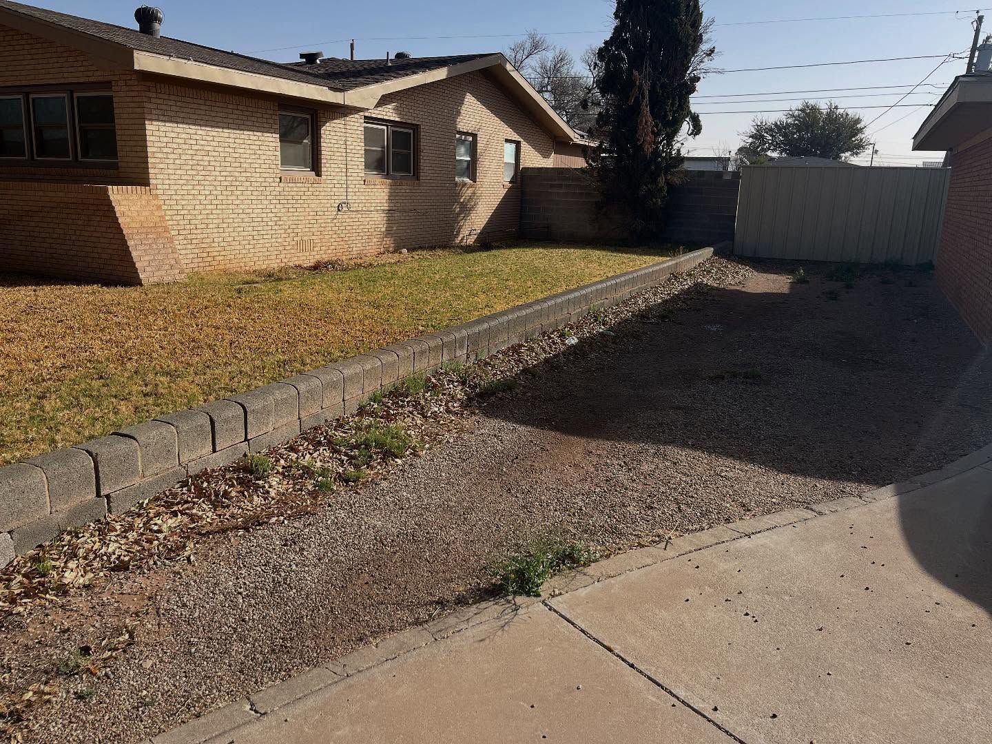 A gravel driveway next to a brick house with a low retaining wall and sparse grass.