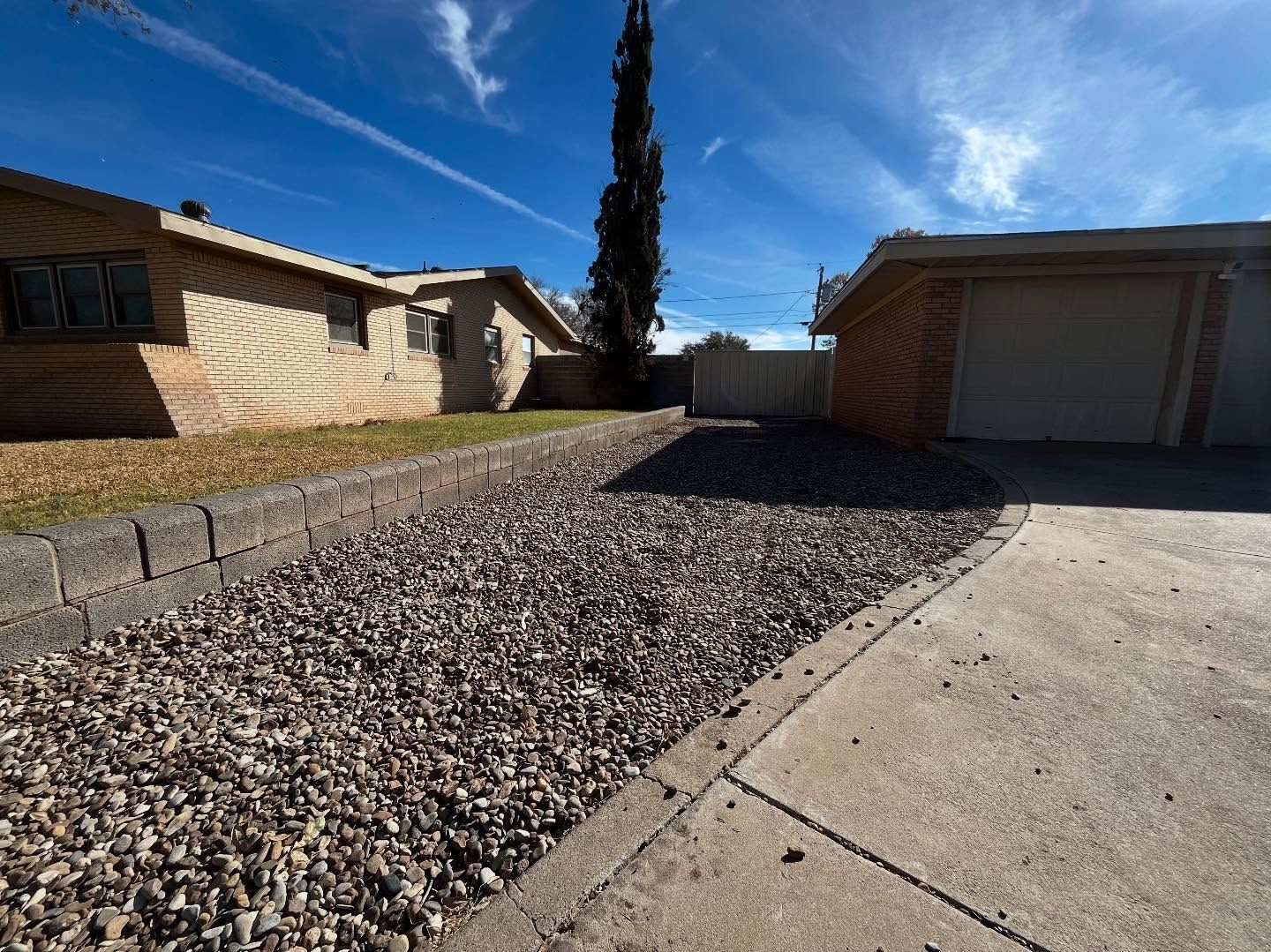 A residential yard with a gravel bed, a brick house, a detached garage, and a tall tree under a blue sky.