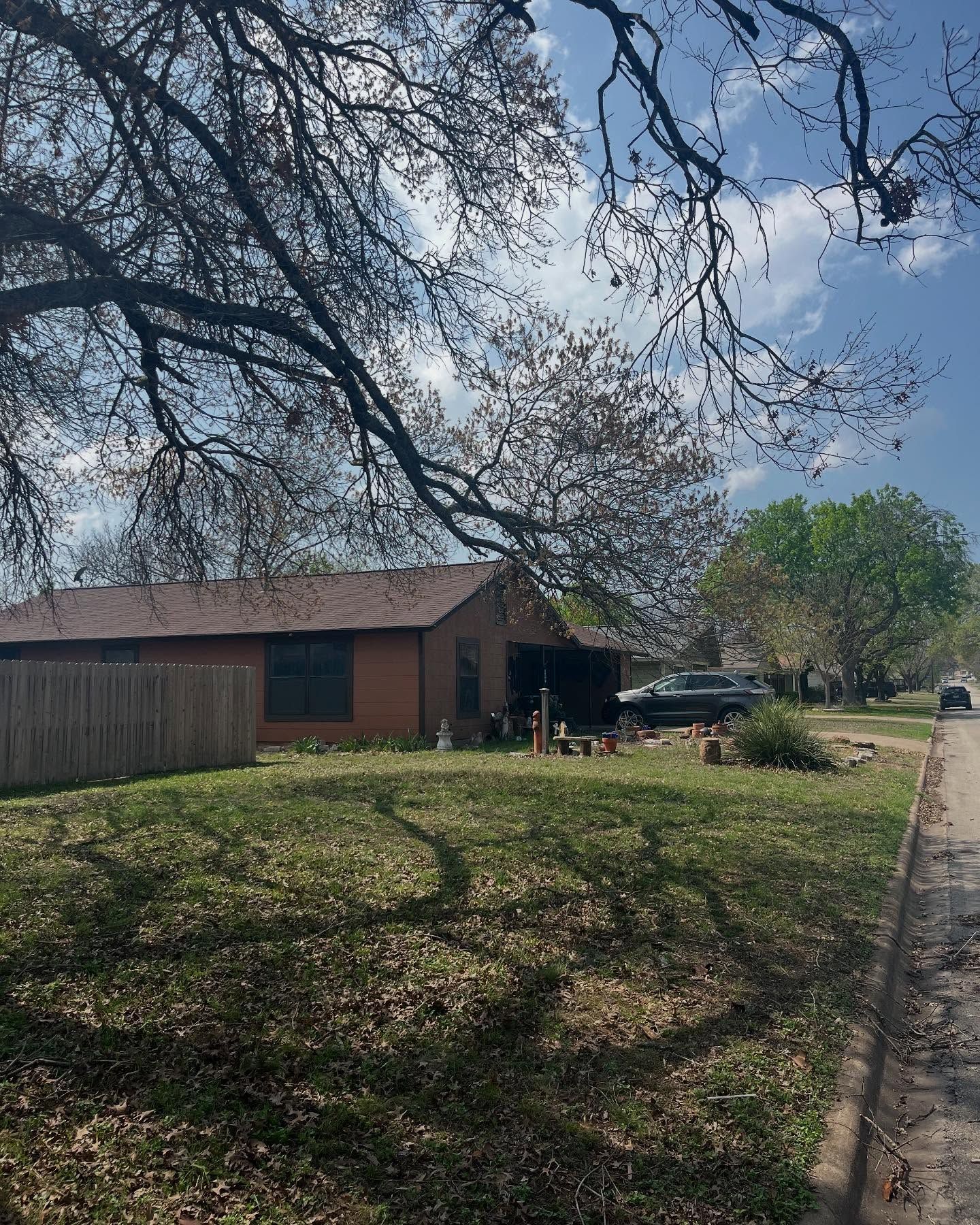 Brick house with brown roof, green lawn, trees, and blue sky. A car sits in the driveway.