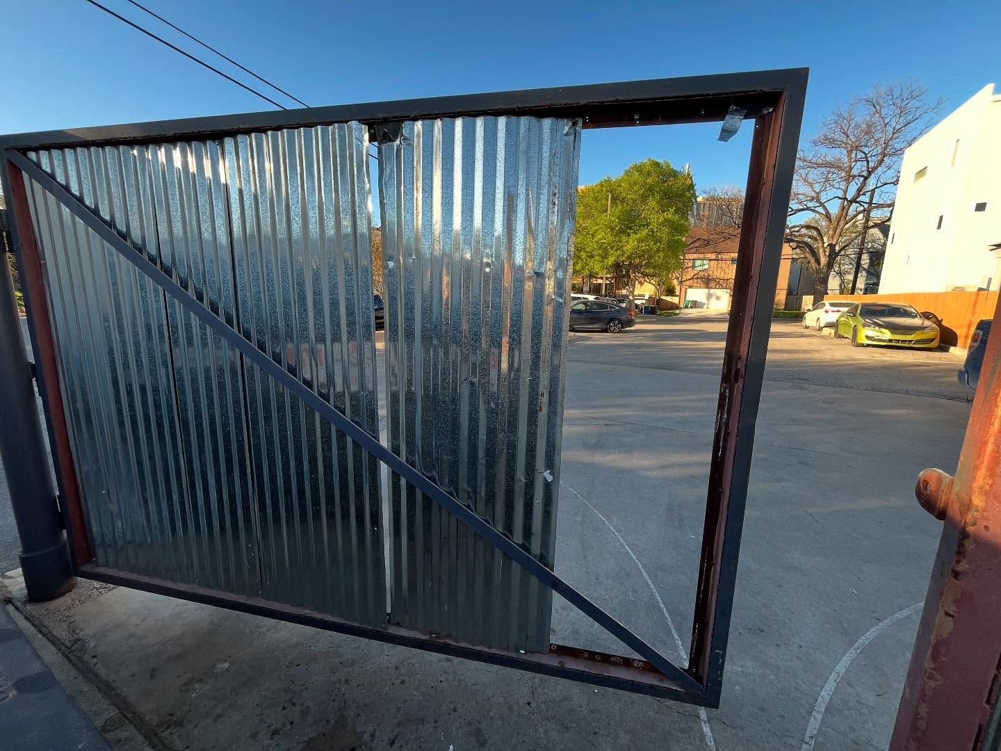 A corrugated metal gate slightly open reveals a parking lot with cars and a building in the background.