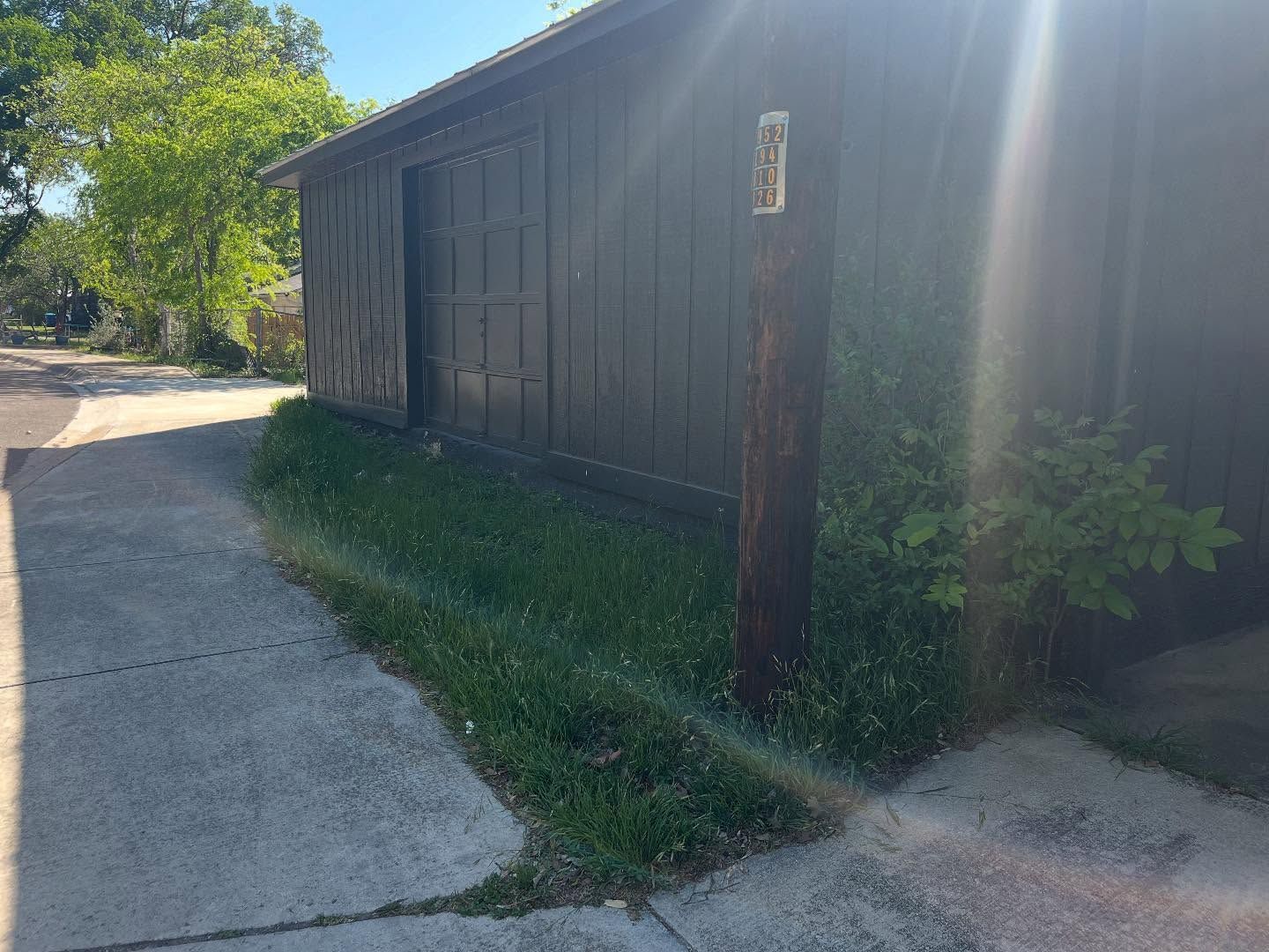 A dark wooden building with a partially open door, beside a sidewalk and a patch of grass.