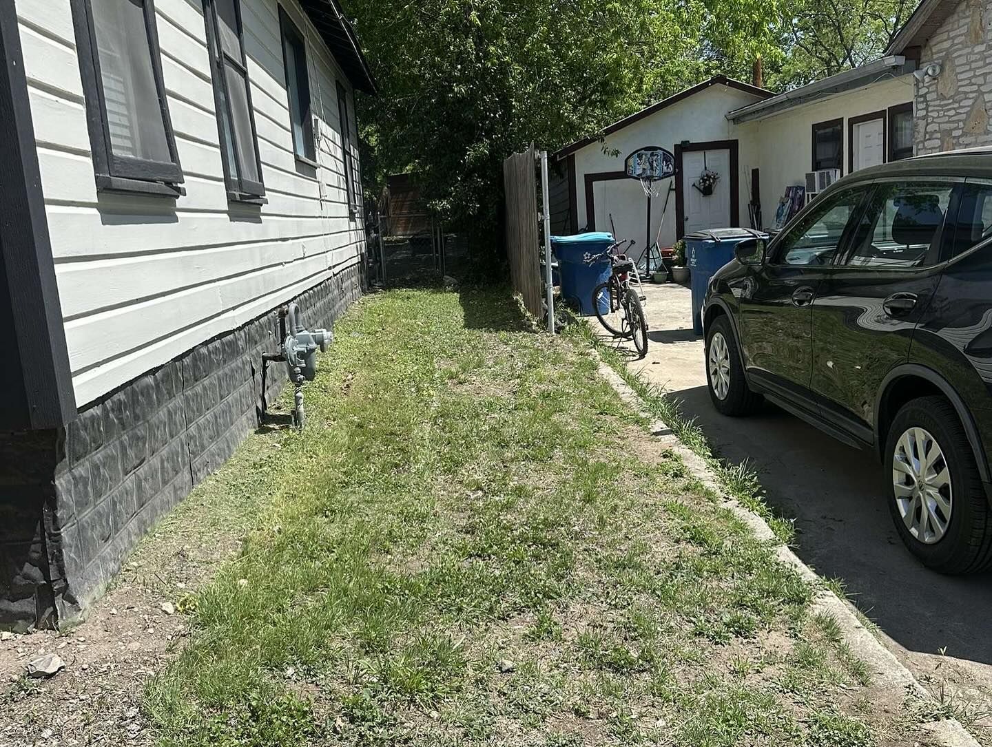 Side view of a house and driveway. A black car is parked next to a narrow grassy area with a bike and trash bins.