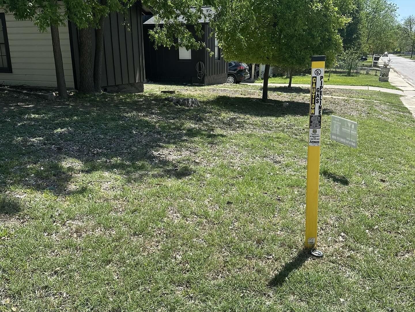 Yellow marker post in grass, likely indicating underground utilities, near a sidewalk and houses.