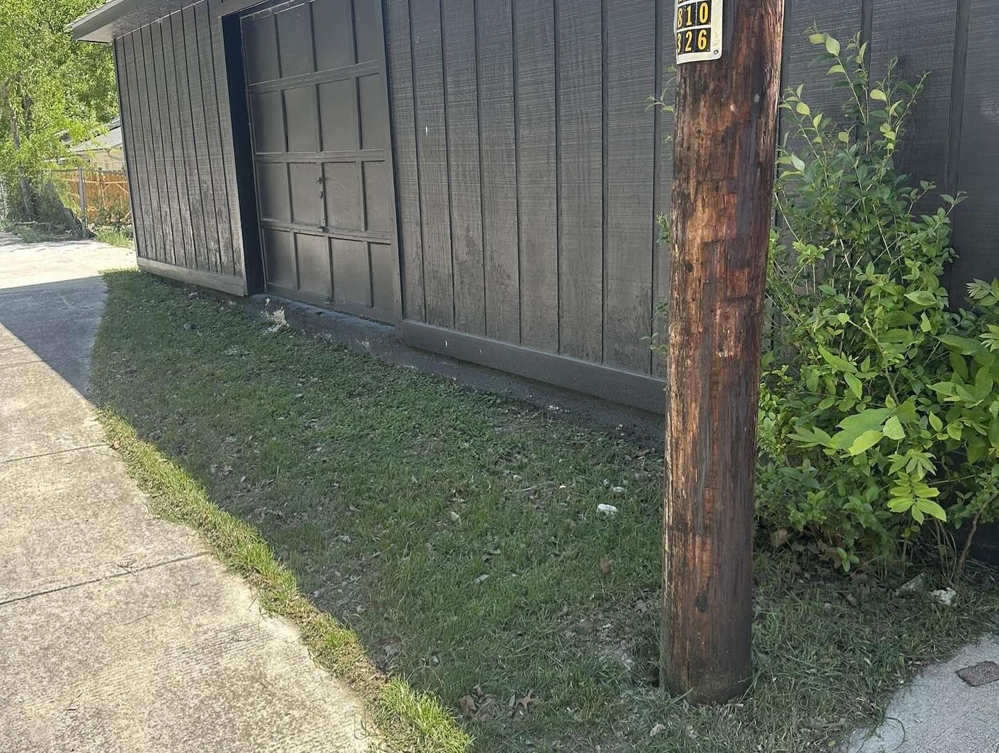Wooden utility pole in front of a dark-colored building and grassy area next to a sidewalk.
