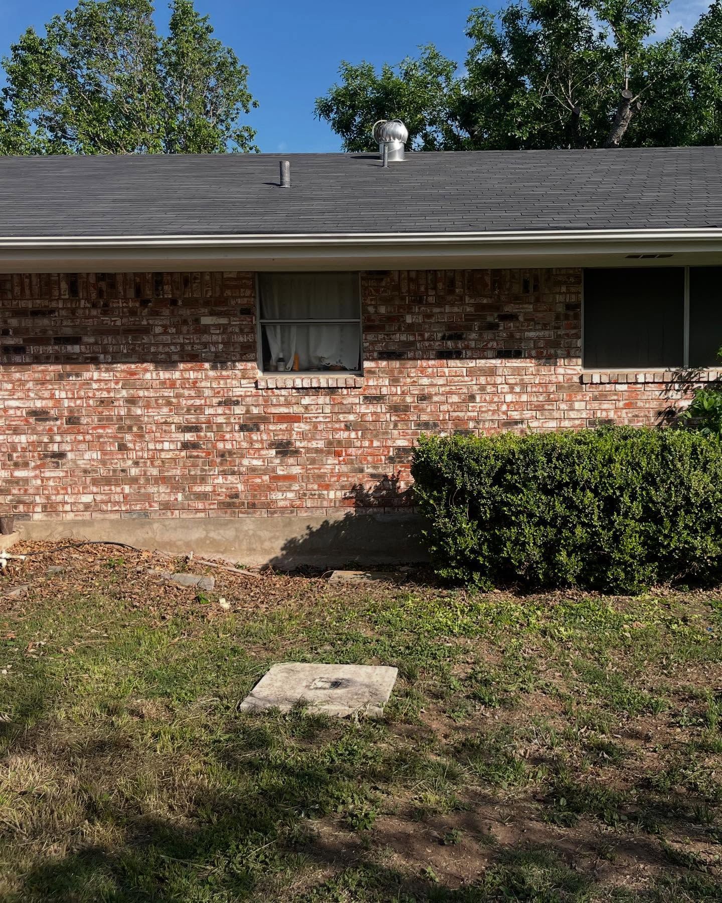 Brick house exterior with a window, bush, and a concrete square on the grass.