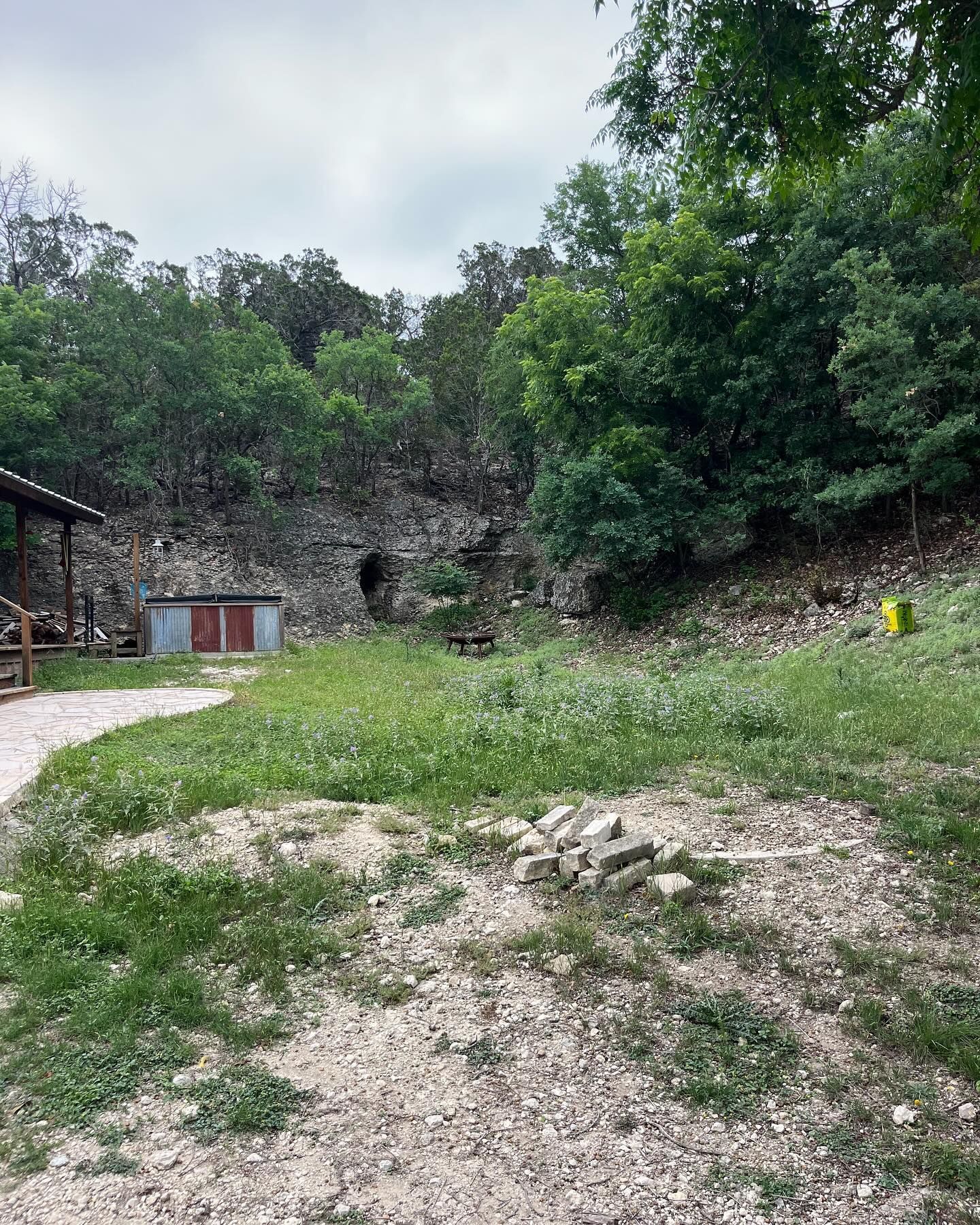 Grassy area with trees, a small shed, and rocky hillside. A dark opening is visible in the rock face.