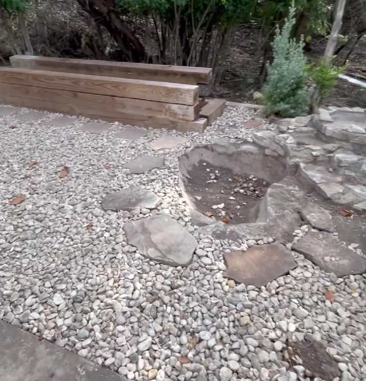Gravel patio with rock border, fire pit, stacked wooden beams, and small tree.