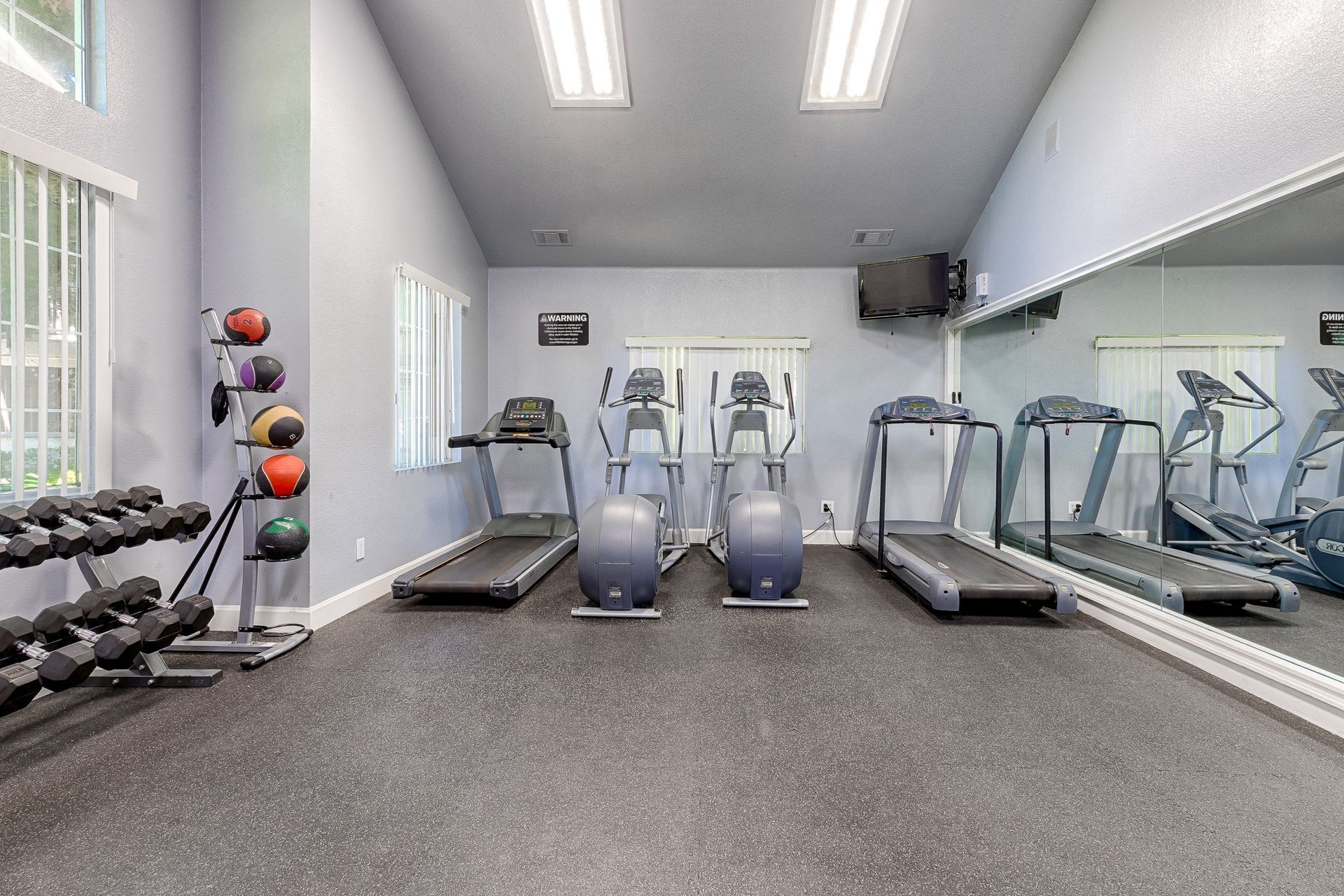 Small gym with treadmills, elliptical machines, free weights, and a mirrored wall.