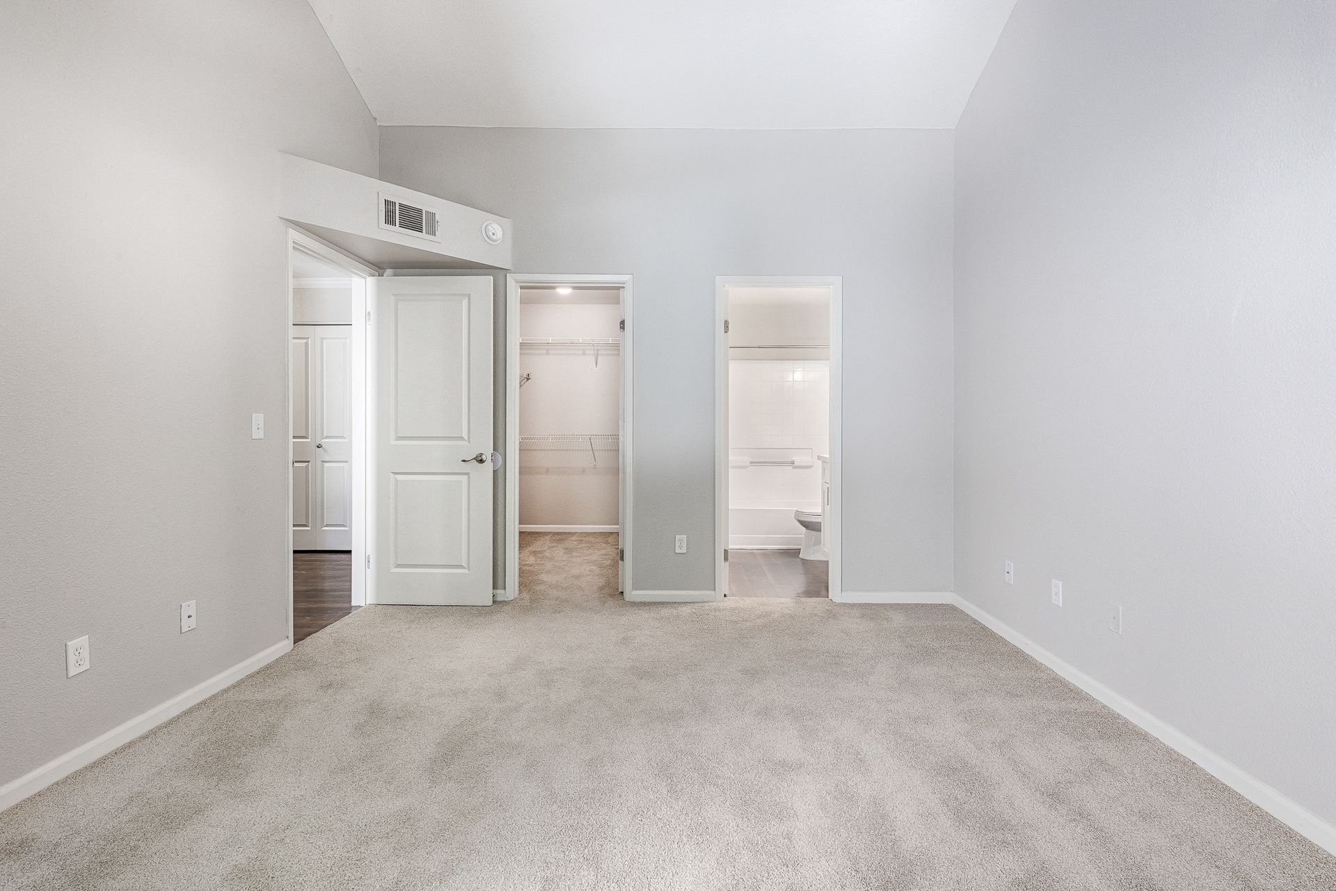 Empty bedroom with light gray walls, beige carpet, and open closet beside a bathroom.