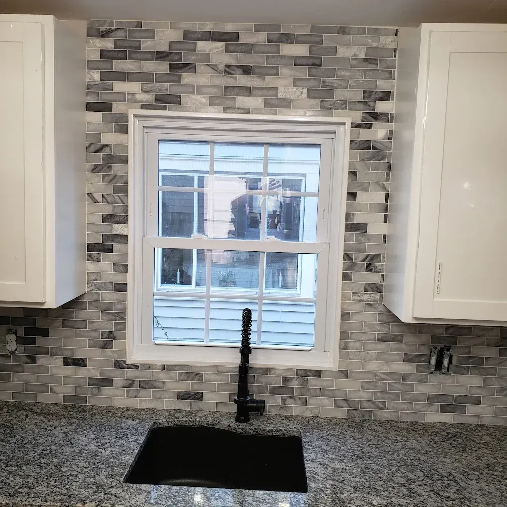 Kitchen backsplash with grey and white mosaic stone tiles, a window, a black faucet, and speckled granite countertops.