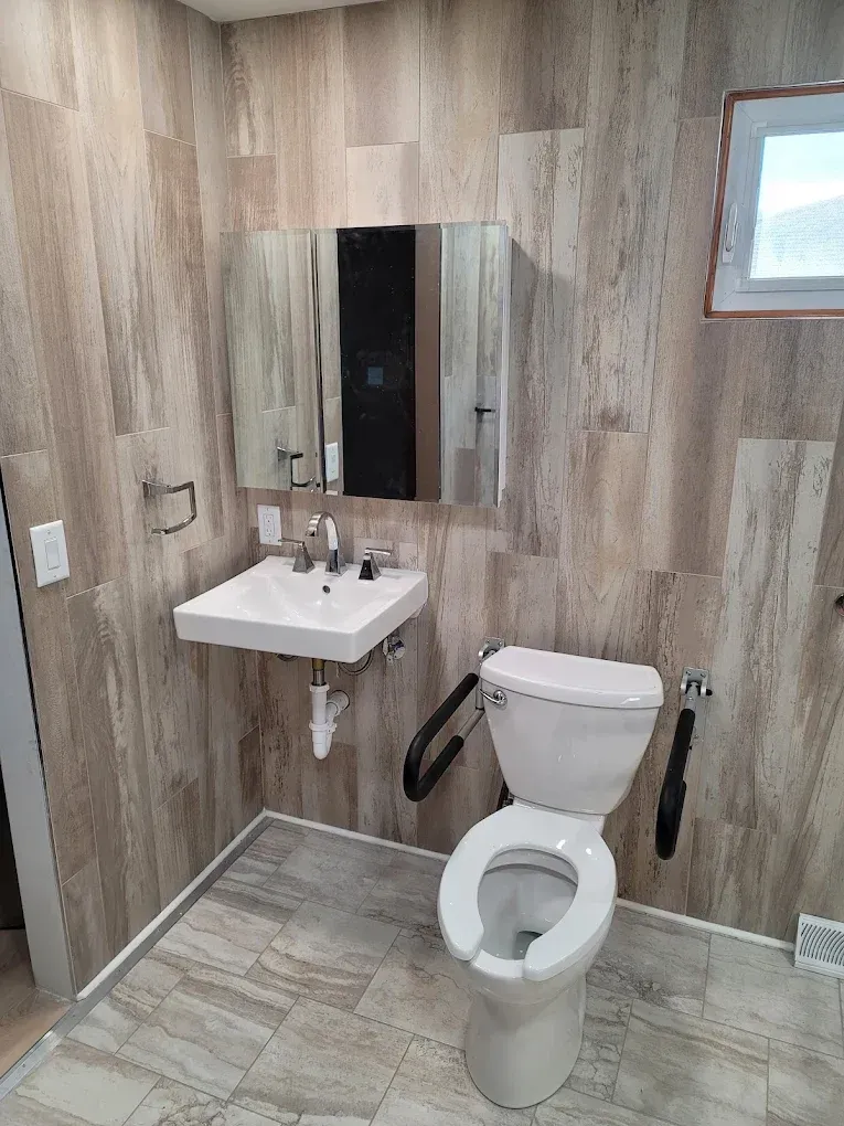 An accessible bathroom with wood-look tile walls, a wall-mounted sink, a toilet with grab bars, and a mirror.