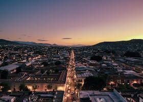Cityscape at dusk, with a long road lined with lights extending into the distance between buildings. Mountains frame the horizon against a purple and orange sky.