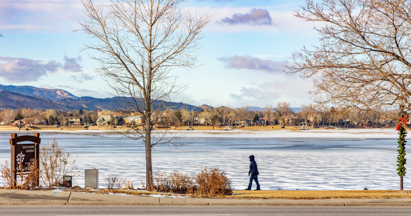 Person walking along a lakefront path, trees, partially frozen water, and mountains in the background.