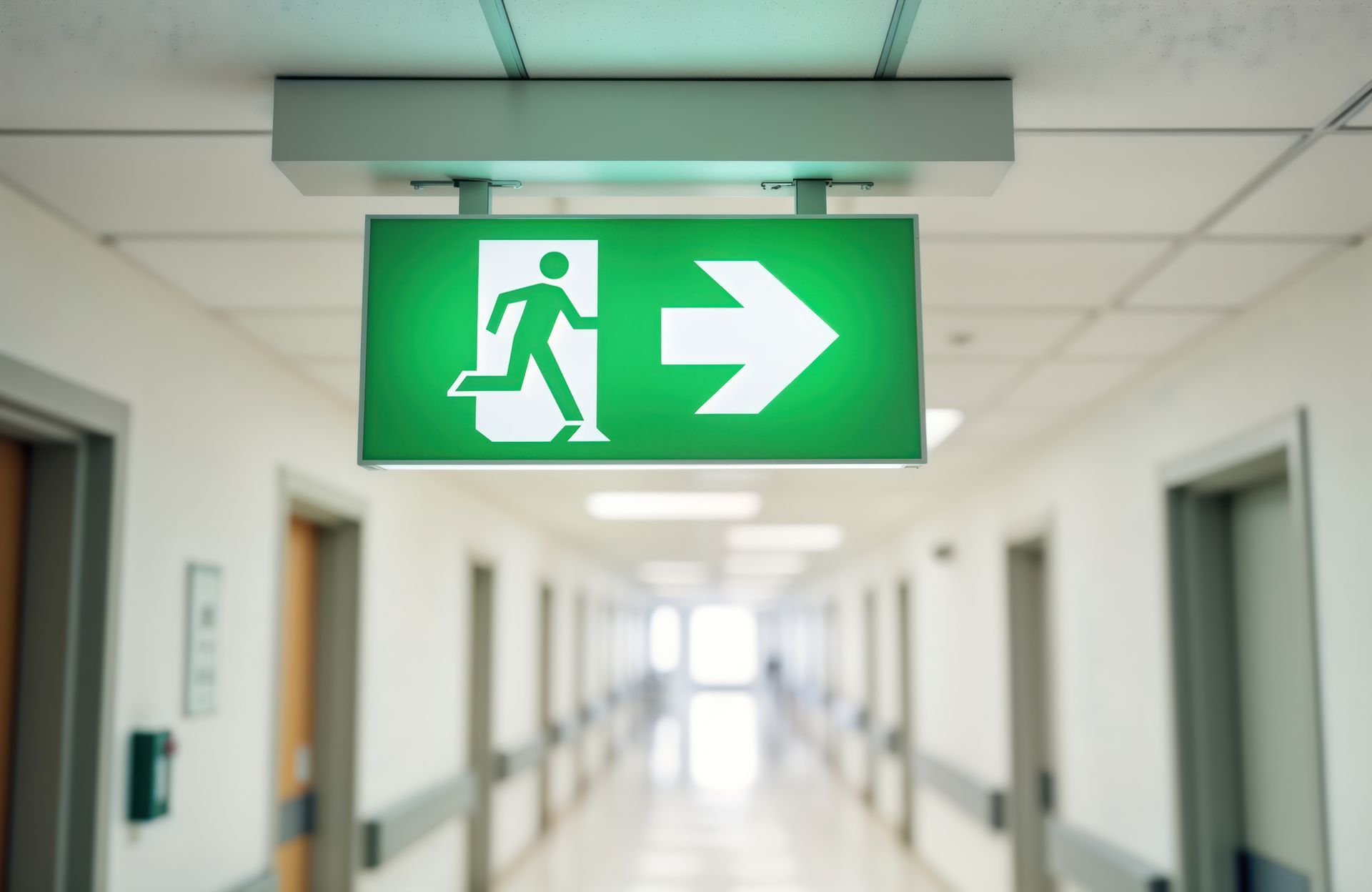 Ein grün beleuchtetes Notausgangsschild über einem Flur, darauf eine nach rechts laufende Figur.
