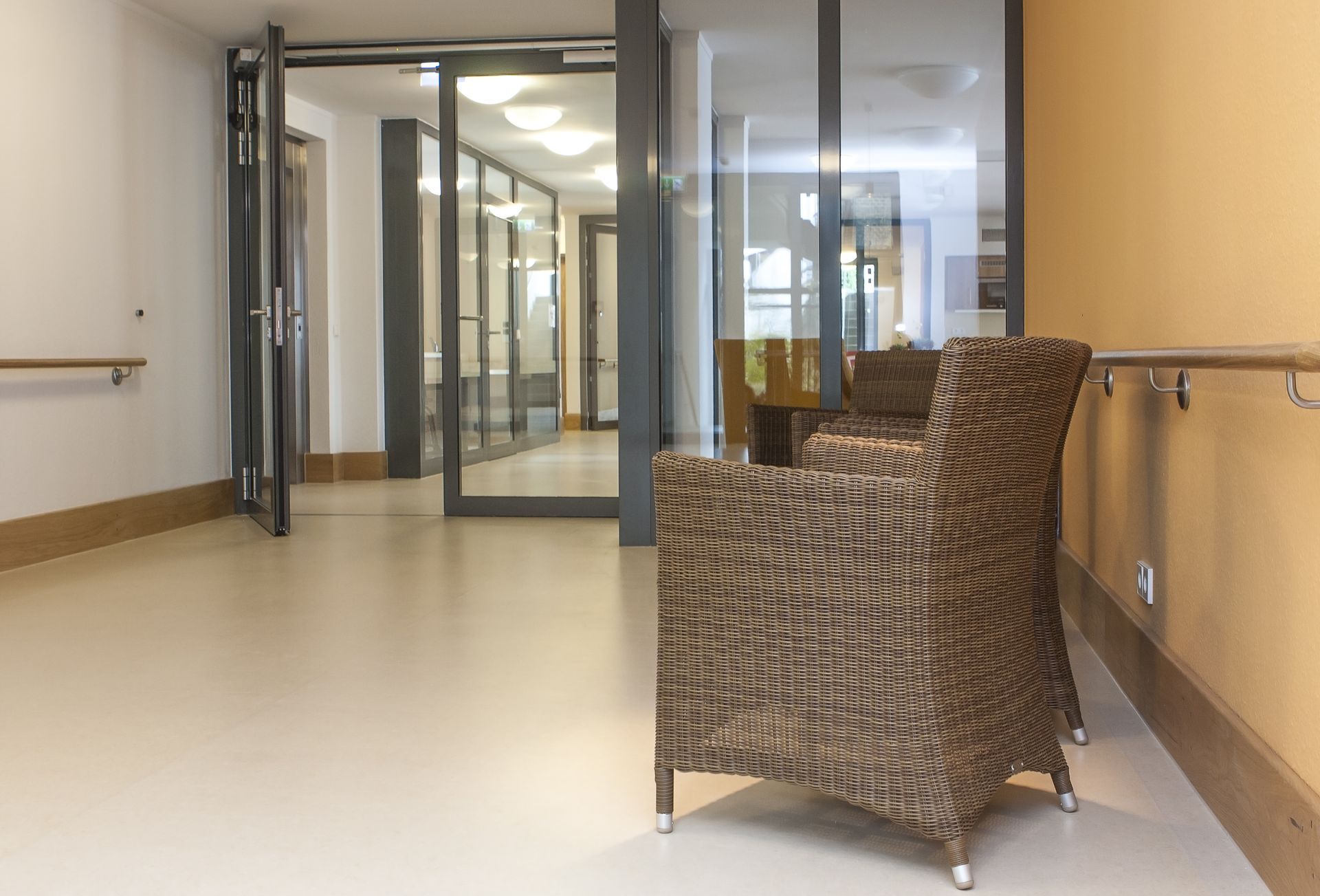 Korbstühle in einem hell erleuchteten Flur. Dahinter sind Glastüren und ein langer Korridor zu sehen.