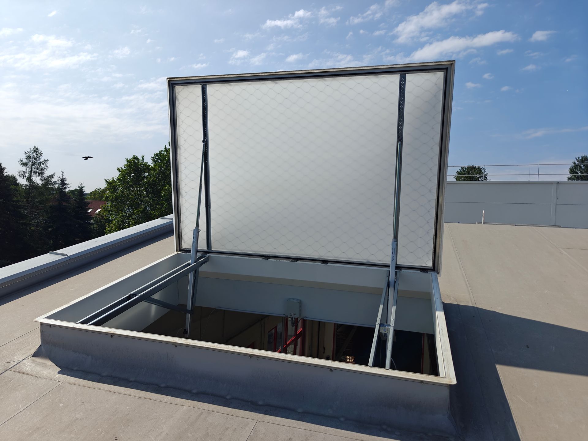Offenes Dachfenster auf einem Flachdach mit durchsichtiger, aufklappbarer Abdeckung. Blauer Himmel im Hintergrund.