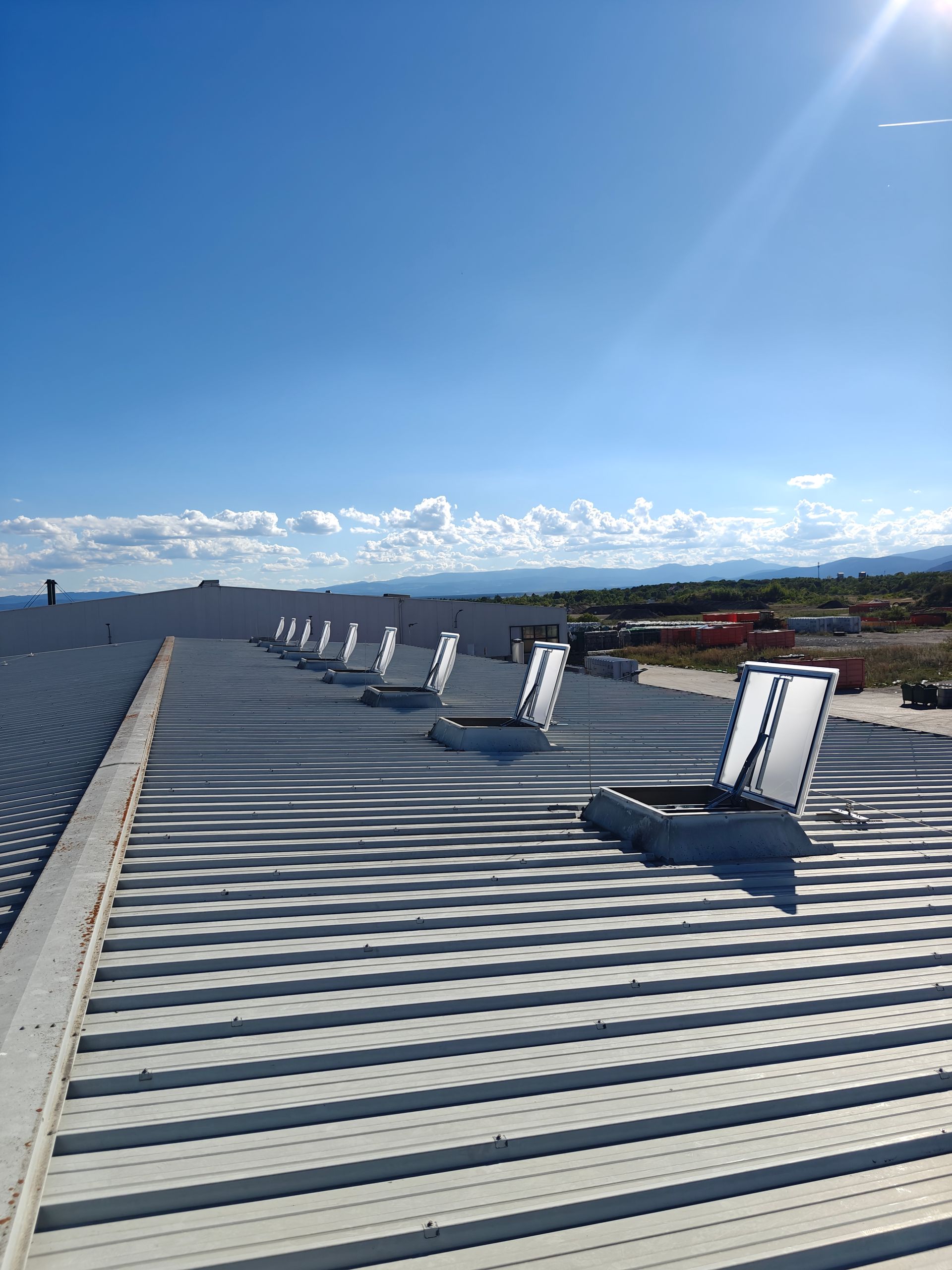 Metalldach mit Oberlichtern unter klarem, strahlend blauem Himmel. Im Hintergrund ferne Berge.