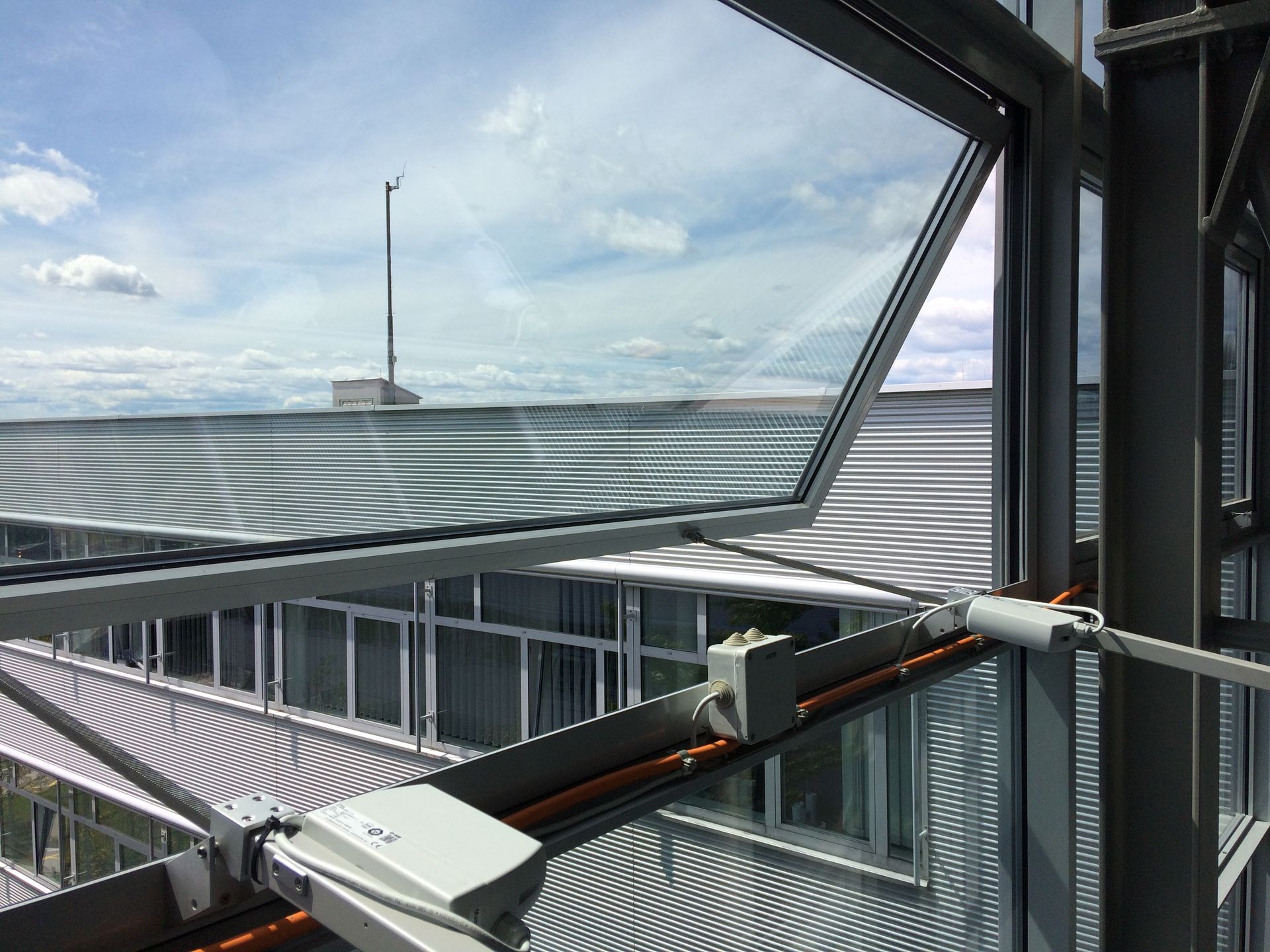 Geöffnetes Fenster mit Blick auf ein Gebäude mit Wellblechdach, einen Mast und einen bewölkten Himmel.