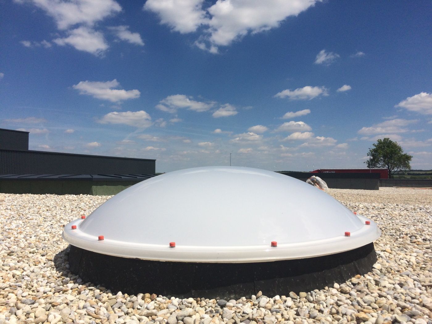 Weiße, kuppelförmige Lichtkuppel auf einem Kiesdach vor blauem Himmel mit Wolken.