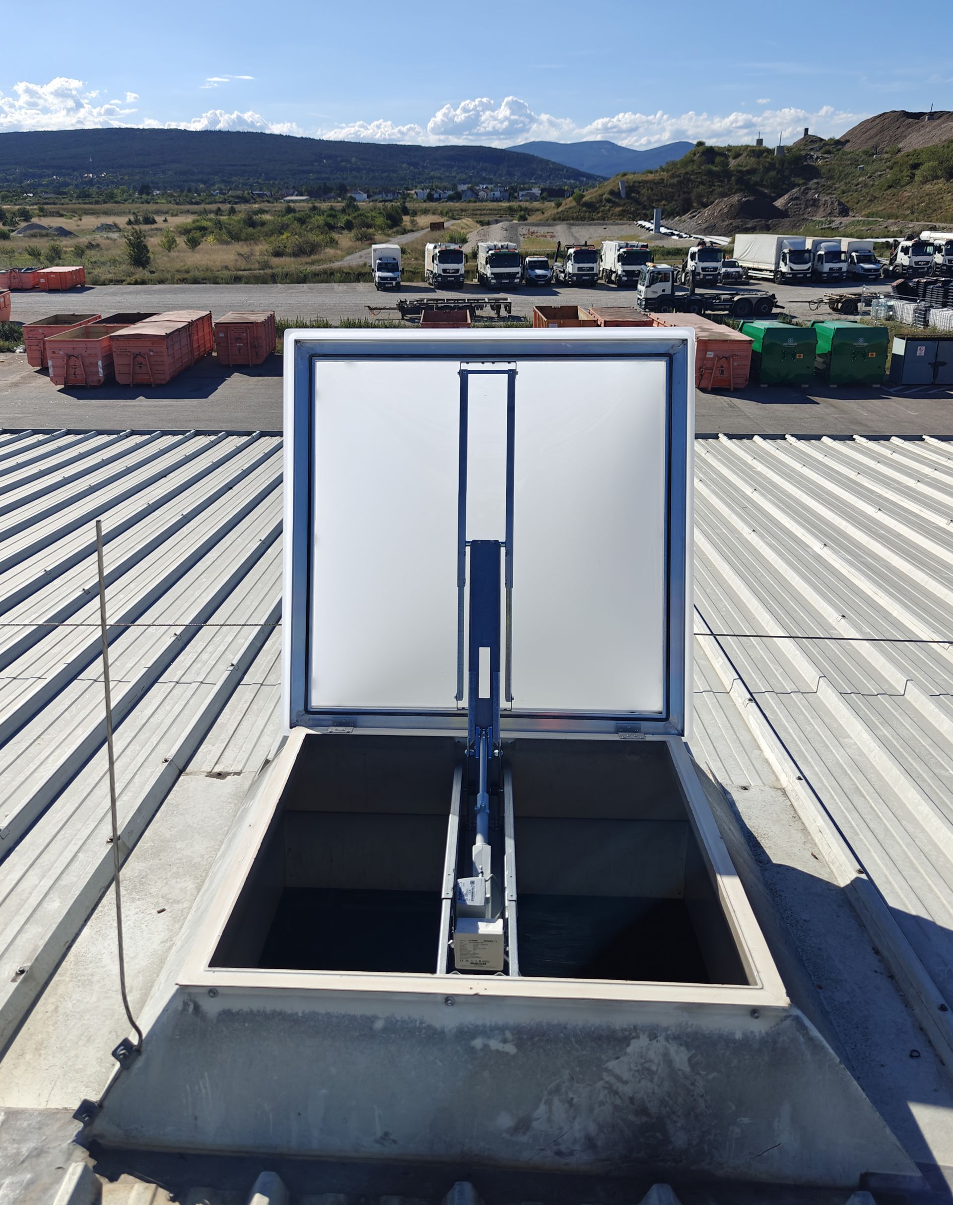 Zugangsluke im Dach, im Hintergrund sind blauer Himmel, Berge und geparkte Fahrzeuge zu sehen.