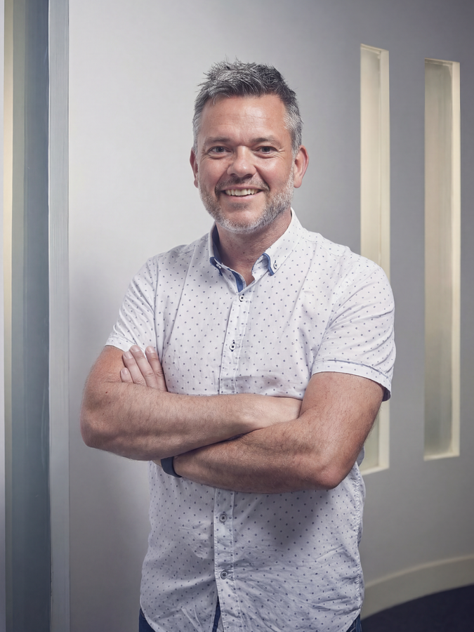 Man with crossed arms in a white dotted shirt, standing near a wall in a bright setting. Managing Director of ACi Studios 