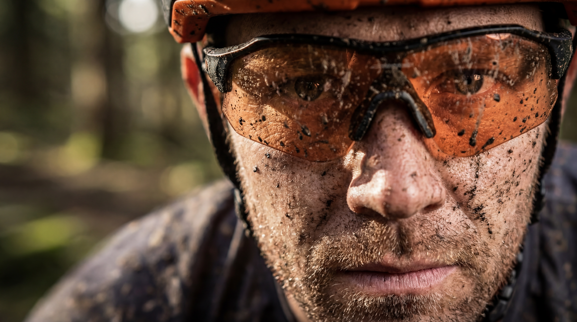 Close-up of a person with mud-covered face and orange-tinted cycling glasses in a forest setting.