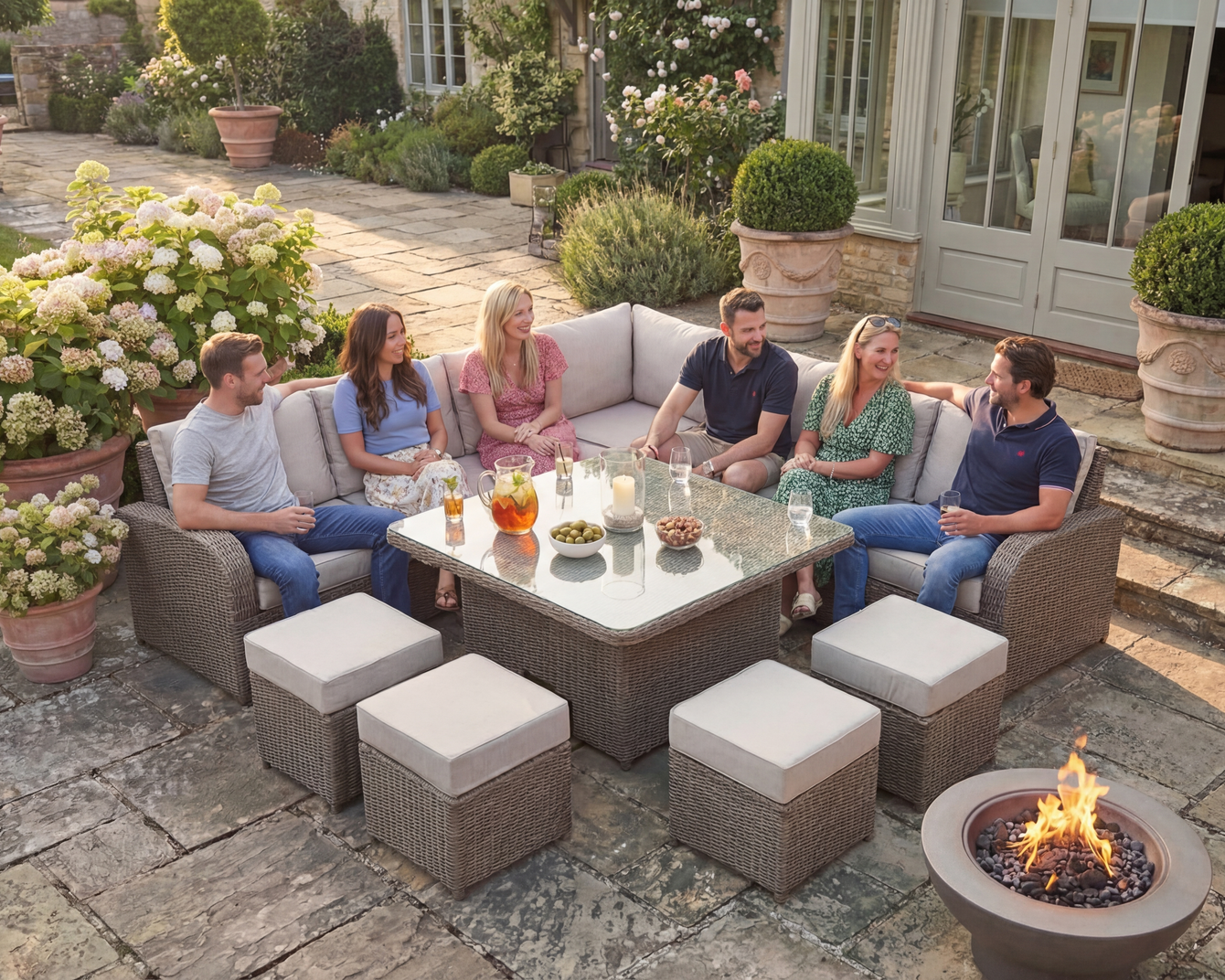 Group of people sitting around outdoor sectional and table; fire pit in foreground.