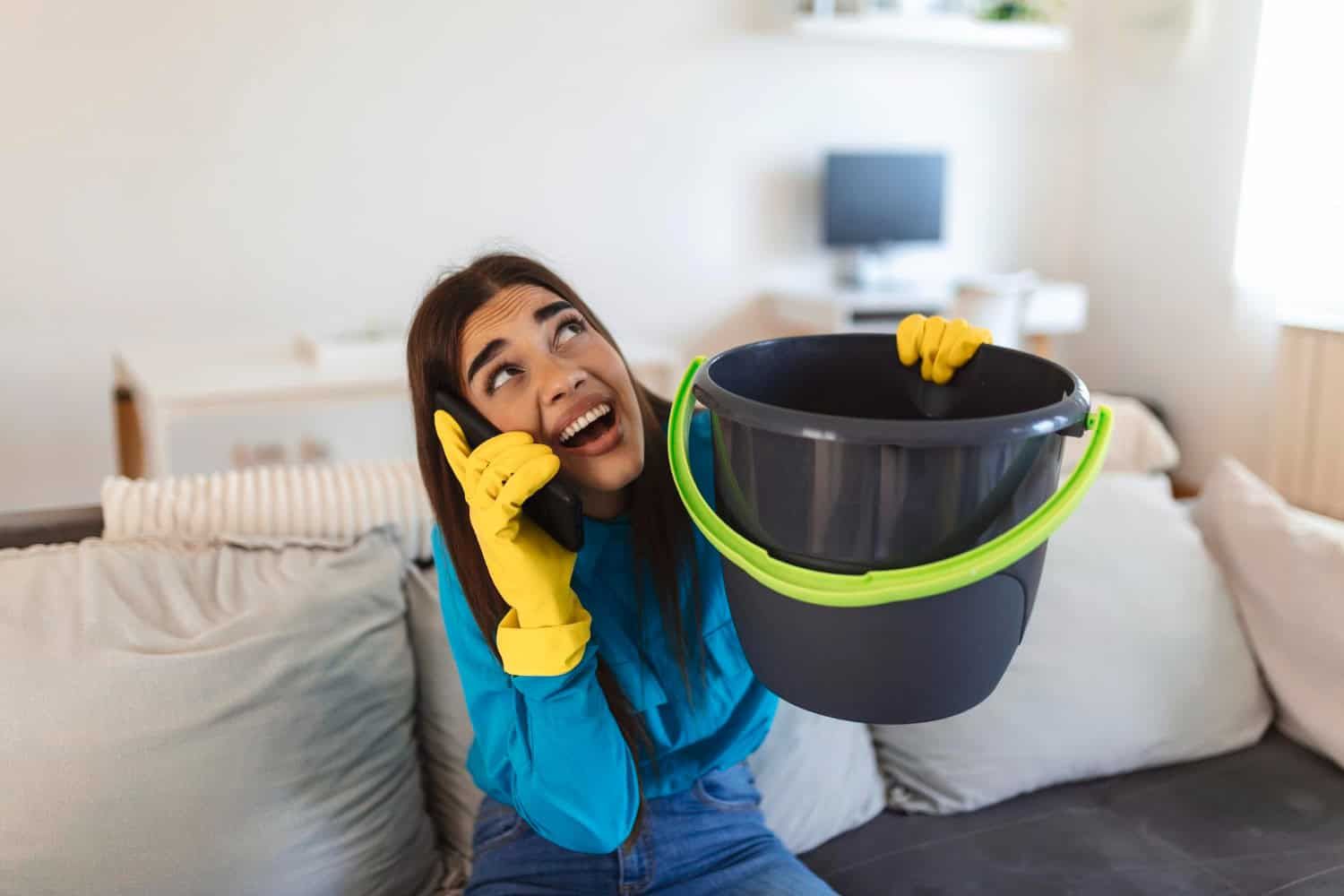 A woman calling plumber because of a burst pipe and holding a bucket to contain a water leak