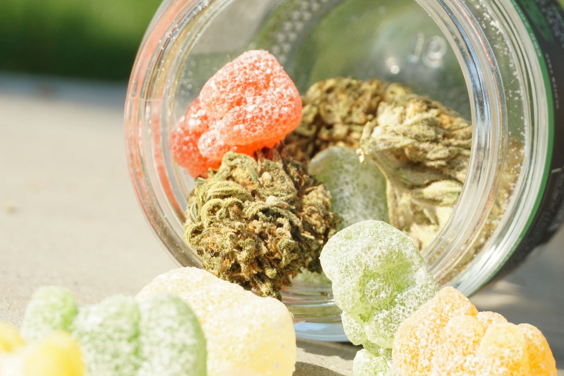 A glass jar tipped over, spilling cannabis buds and colorful sugar-coated cannabis-infused gummies onto a surface.