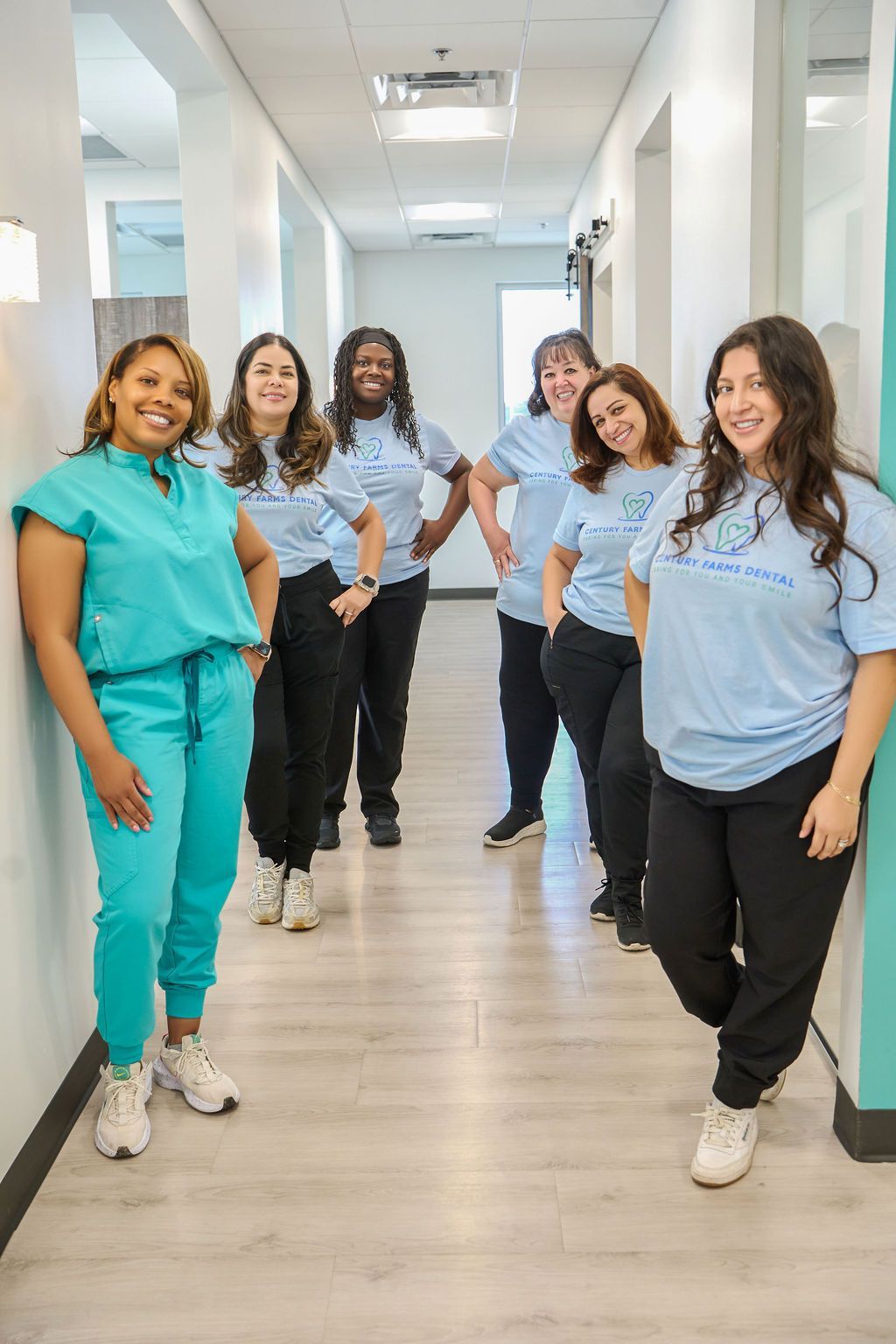 A group of people wearing light blue shirts and black pants smile in a hallway.