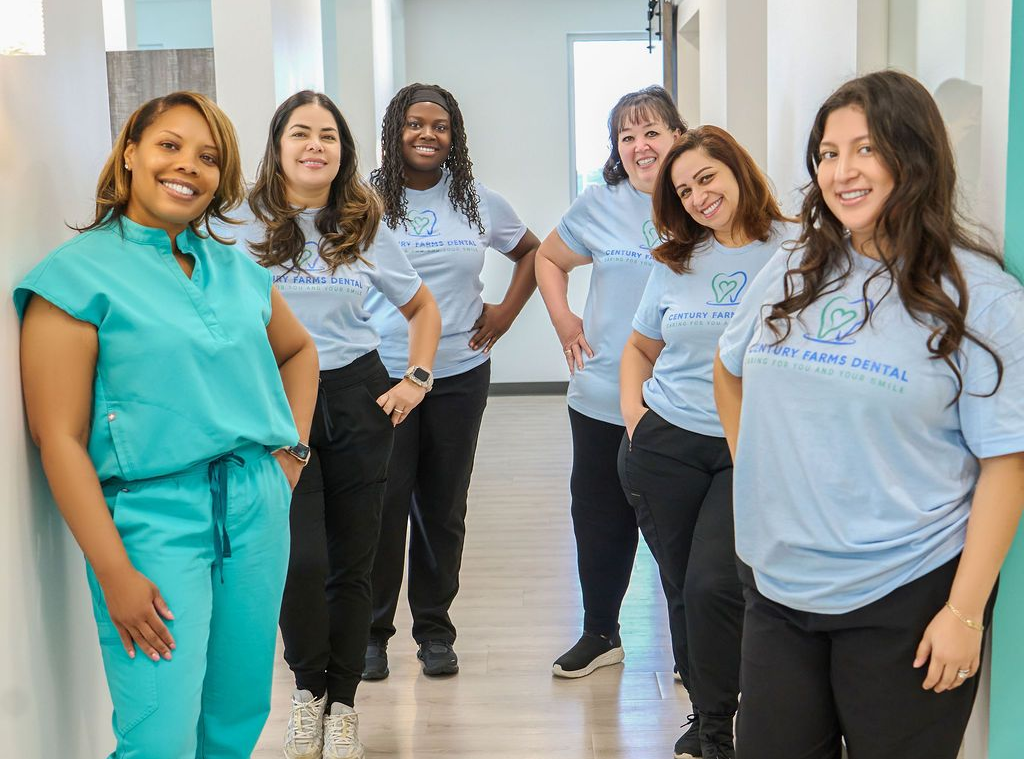 Seven healthcare workers pose in a hallway; some wear blue shirts, black pants, and scrubs; smiling.