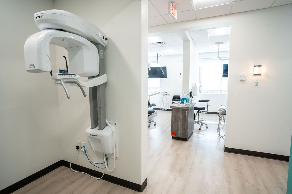 Dental X-ray machine mounted on a white wall in a dental office, with a view into the treatment area.