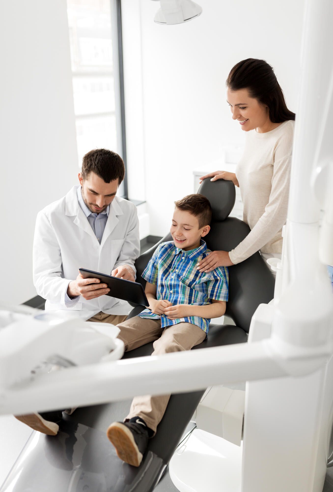 A young boy is sitting in a dental chair with his mother while a dentist looks at a tablet.