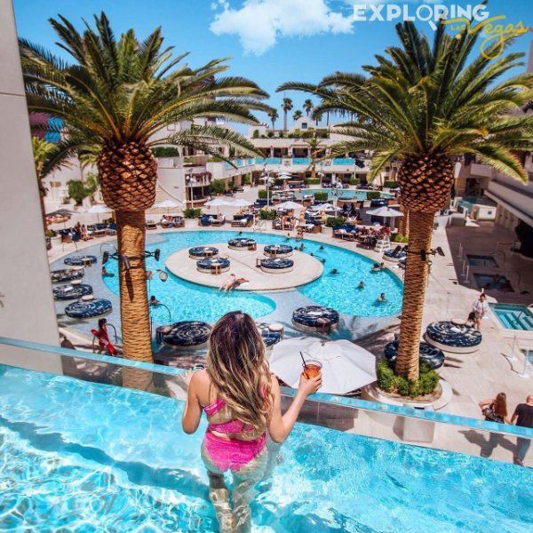 A person in a bright pink swimsuit stands in an infinity pool overlooking a resort courtyard with a large blue pool.